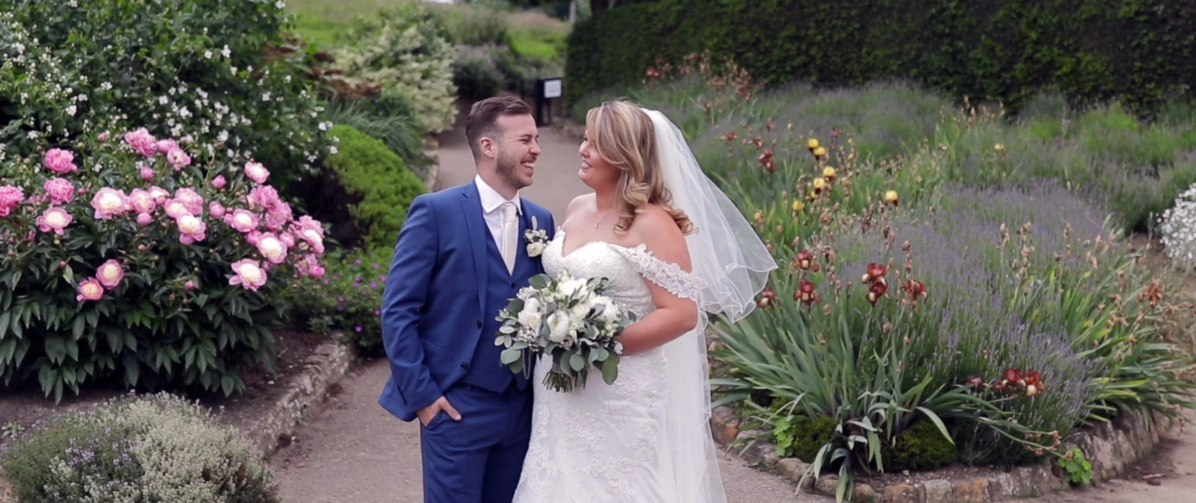 A newlywed couple sharing a joyful moment in a lush, colorful garden. The groom in a blue suit and the bride in a white lace wedding dress hold a bouquet of white flowers, smiling at each other surrounded by vibrant flowers and greenery.