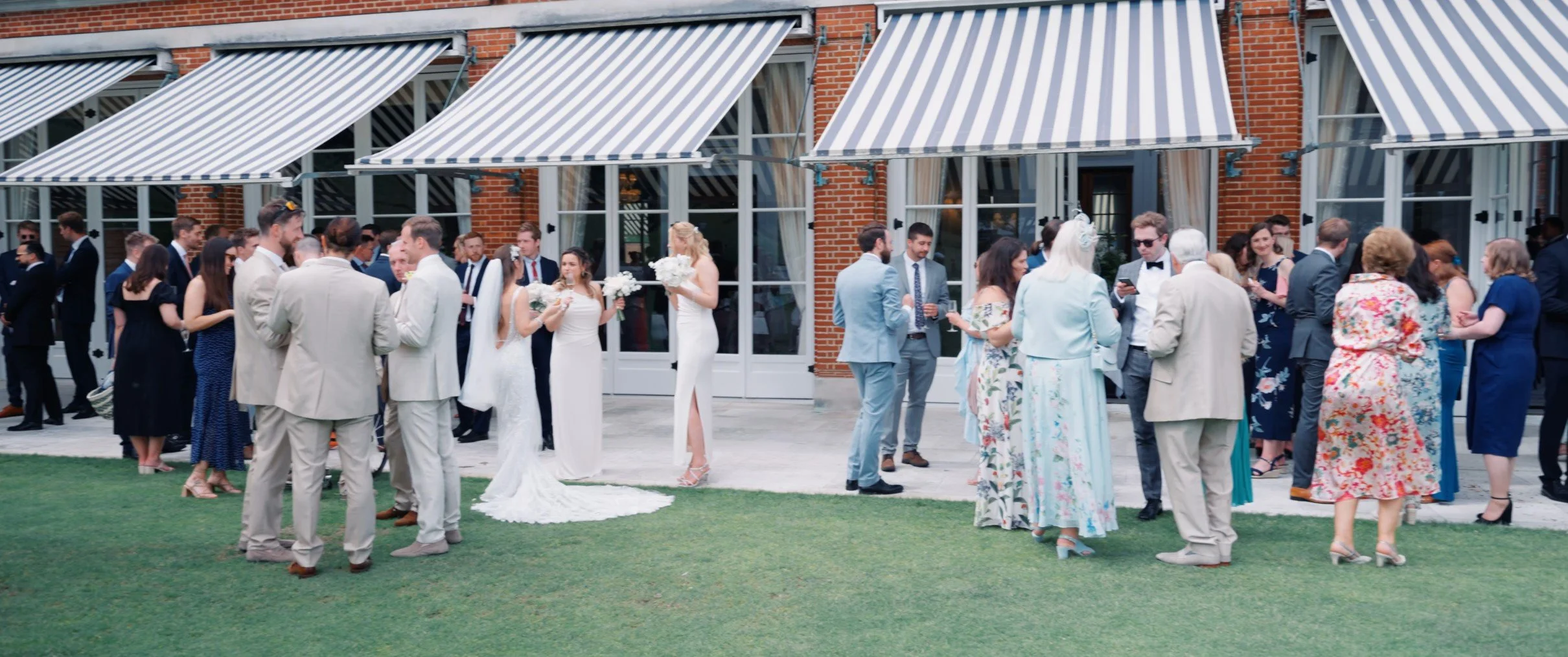 Group of people at a wedding reception outside a brick building with striped awnings, some in formal attire, chatting and taking photos.