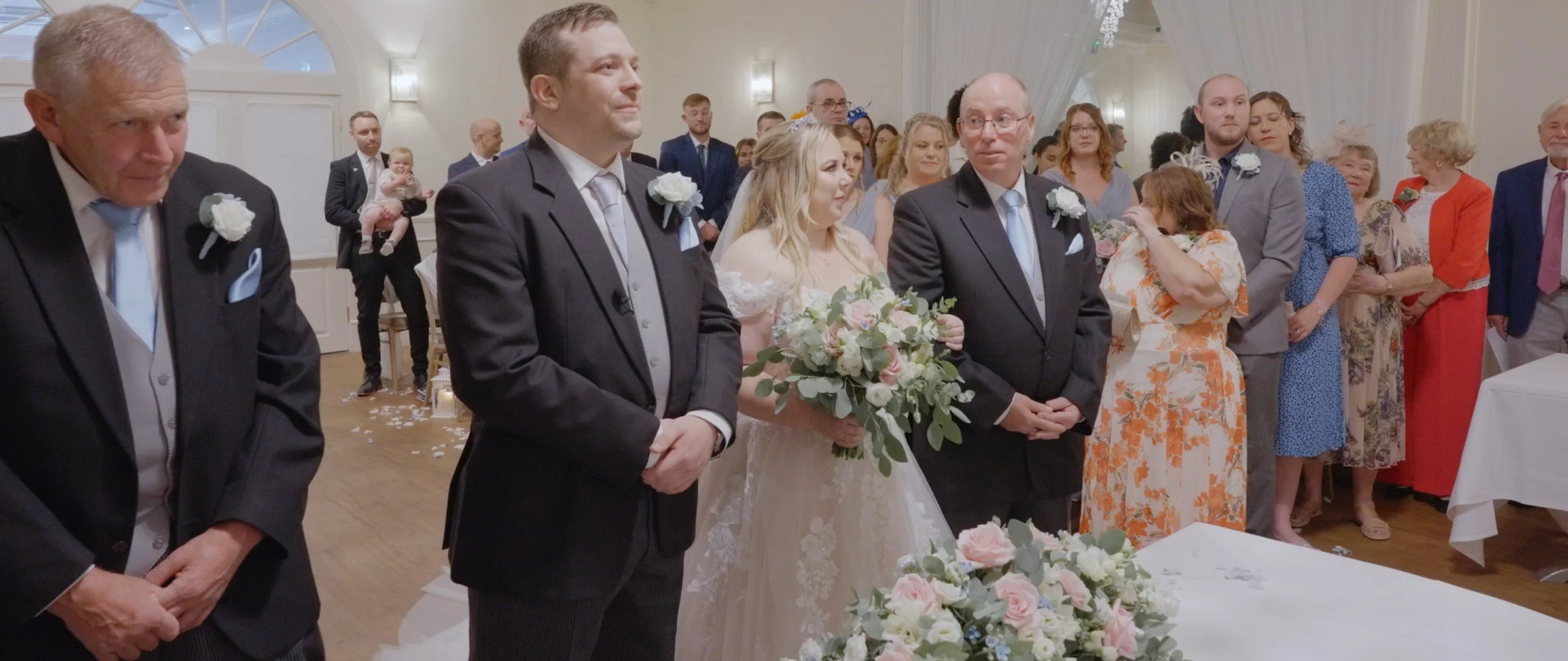Wedding ceremony with bride in a white dress holding a bouquet of pink and white flowers, flanked by groom and family members, surrounded by guests in formal attire in a decorated wedding venue.