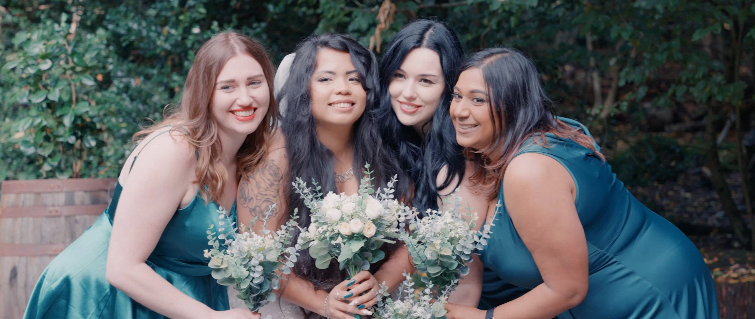 Four women in blue dresses holding bouquets of white flowers, smiling outdoors with green foliage in the background.