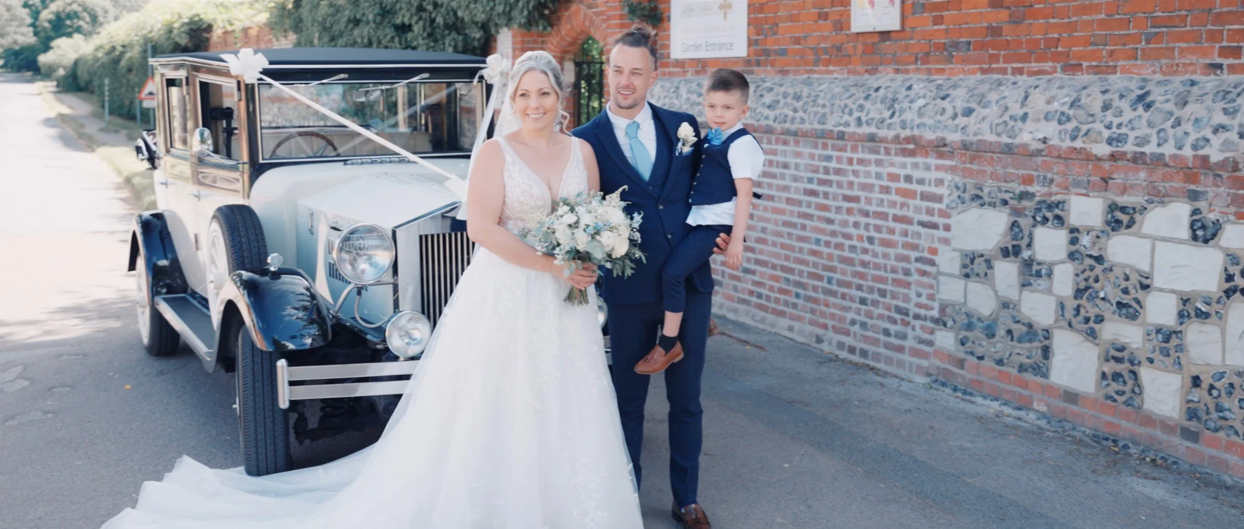 A bride and groom standing next to a vintage car, with a young boy in formal attire, outside a brick and stone building.