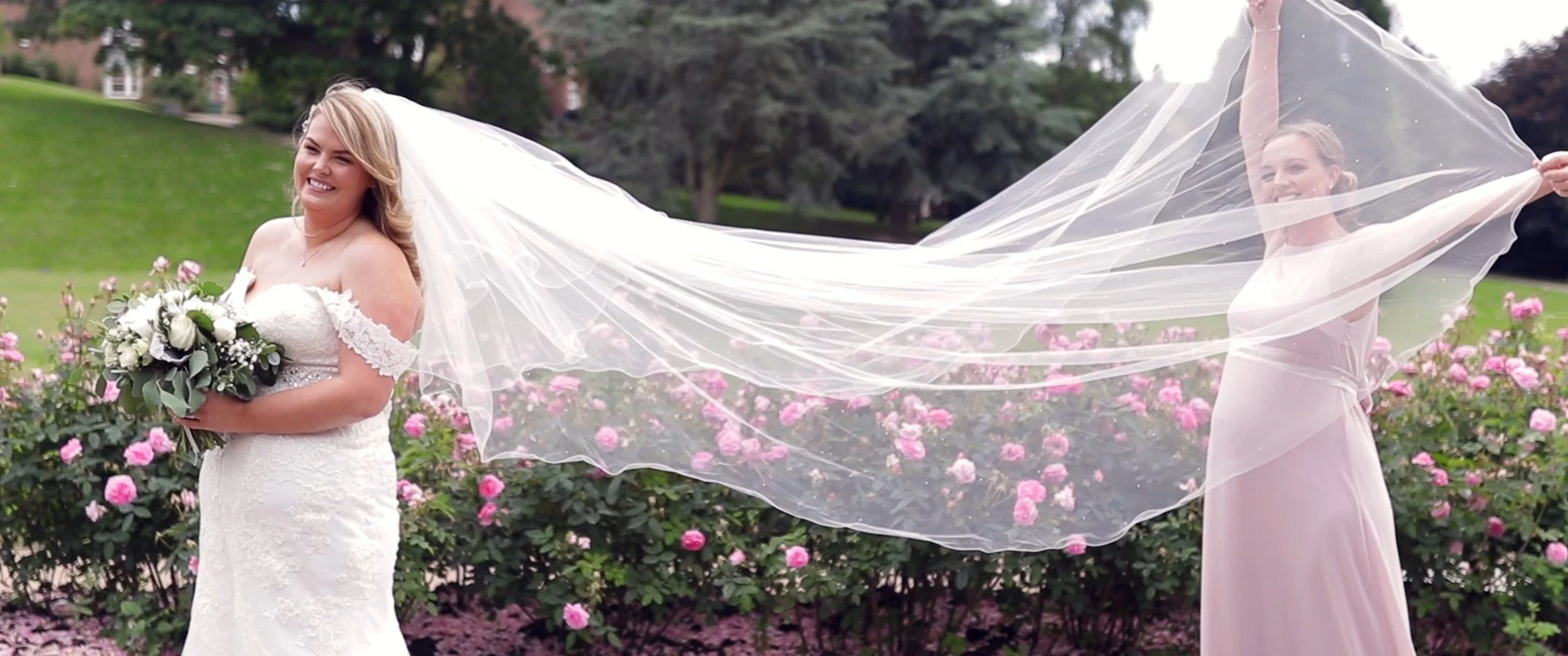 A bride in a white wedding dress holding a bouquet of white flowers, standing outdoors in a garden with pink roses and trees in the background. A woman in a light pink dress is holding part of the bride's veil, which is flowing behind them, creating 