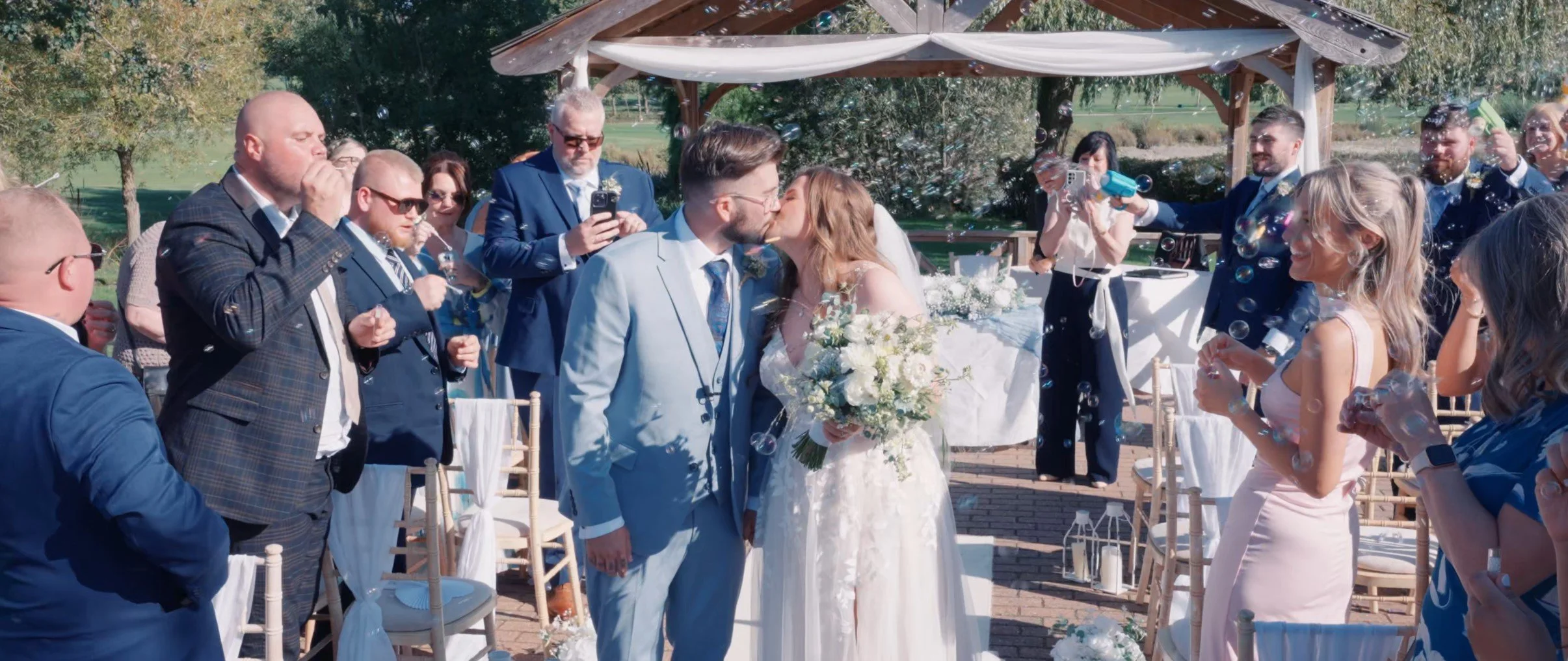 A bride and groom kiss at an outdoor wedding ceremony with friends and family blowing soap bubbles while celebrating under a gazebo.
