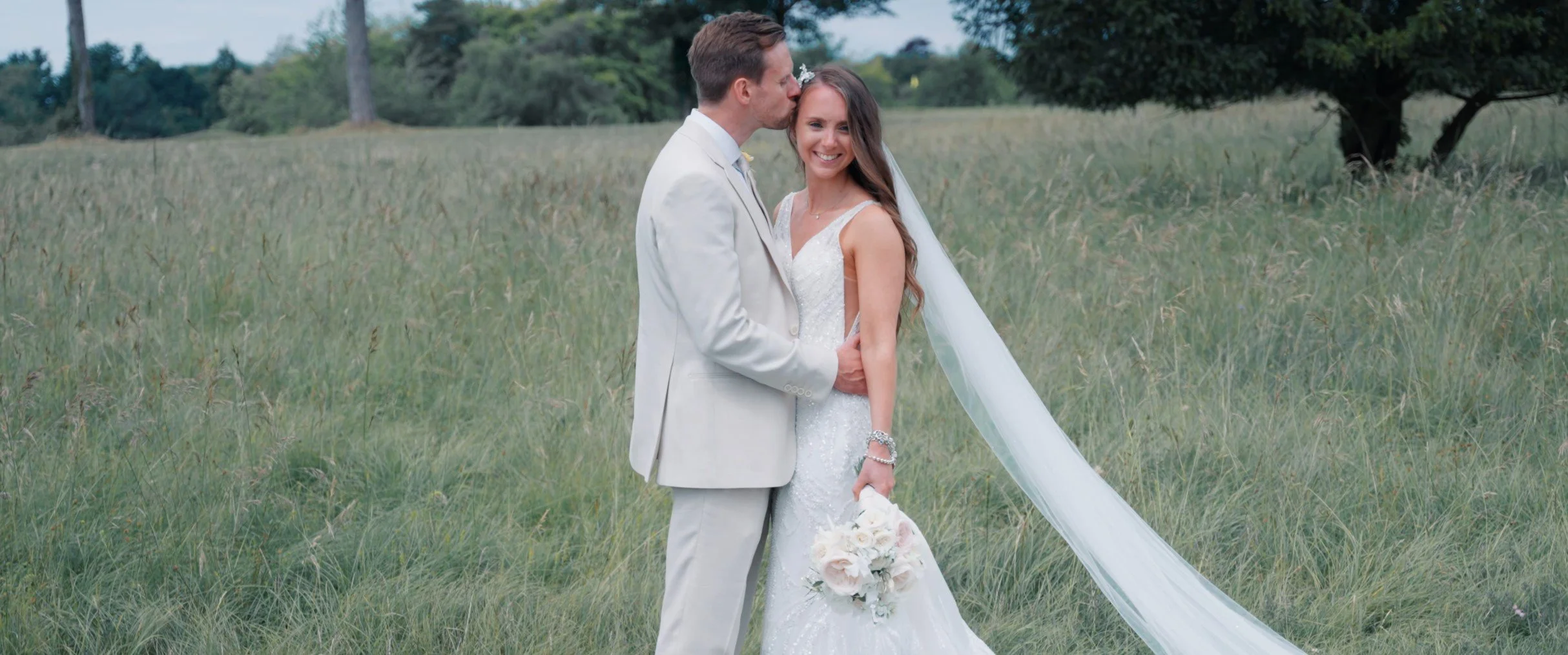 A bride and groom standing in a grassy field, with the groom kissing the bride on her forehead. The bride is smiling and holding a bouquet of white flowers; she is wearing a white wedding dress and veil. The groom is dressed in a light-colored suit.