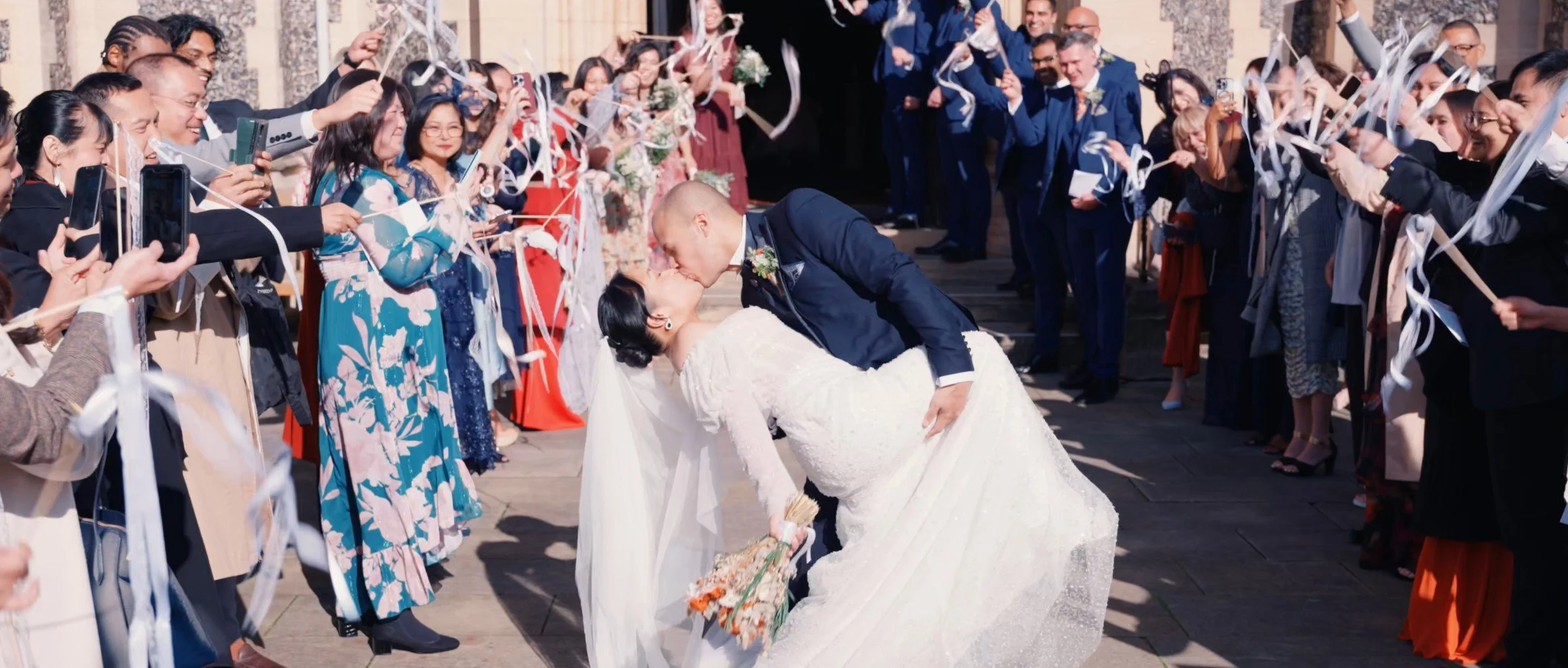 Bride and groom kissing as guests celebrate with confetti and ribbons at their wedding reception outdoors.