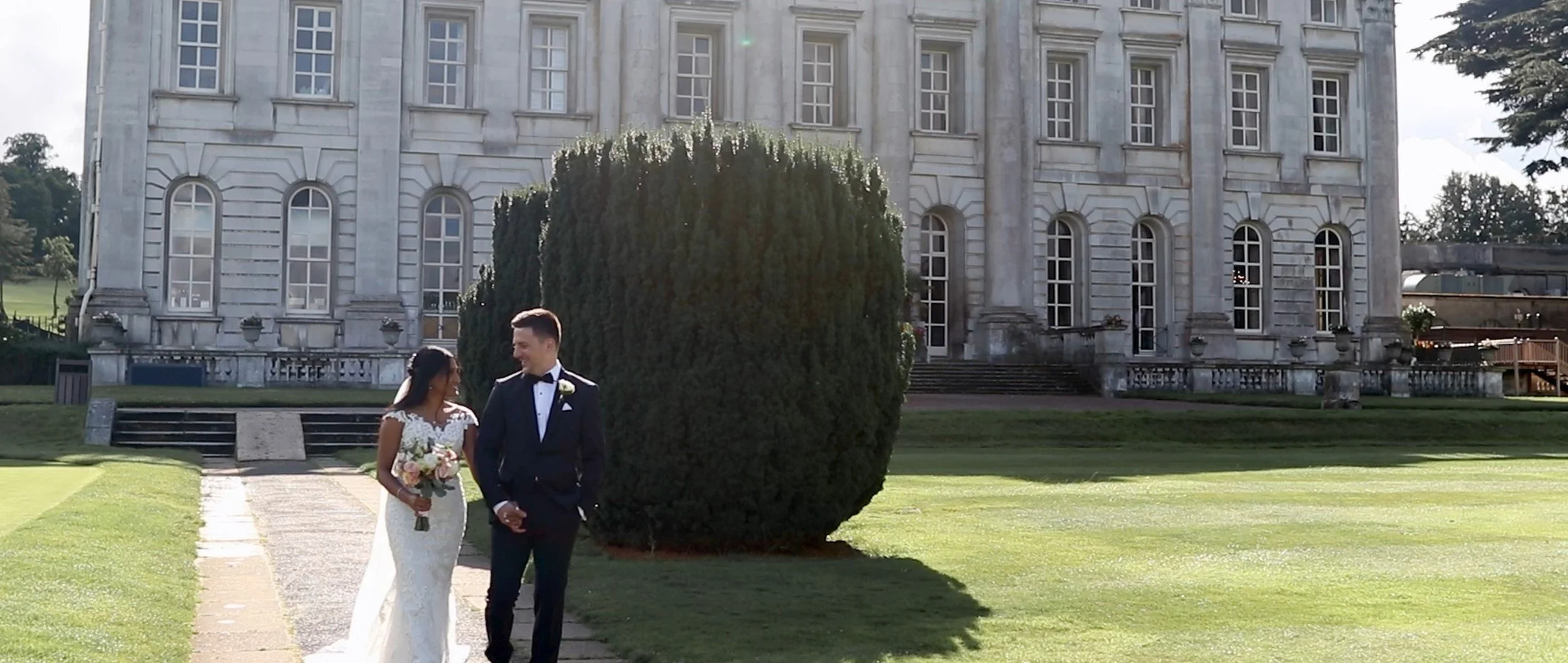 A bride and groom walking on a pathway in front of a large colonial-style mansion with a manicured lawn and round shrub in the background.
