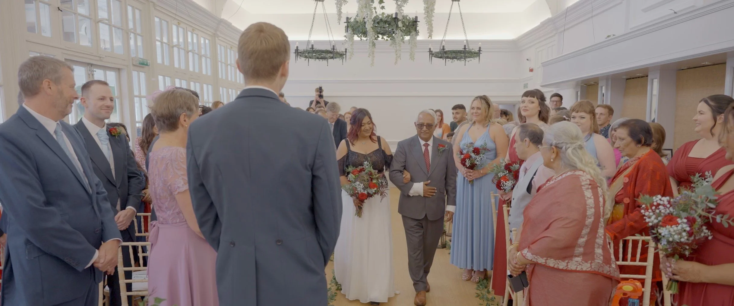 A group of people at a wedding ceremony inside a bright, elegant venue with white walls and large windows, standing in a semicircle around the bride and groom.