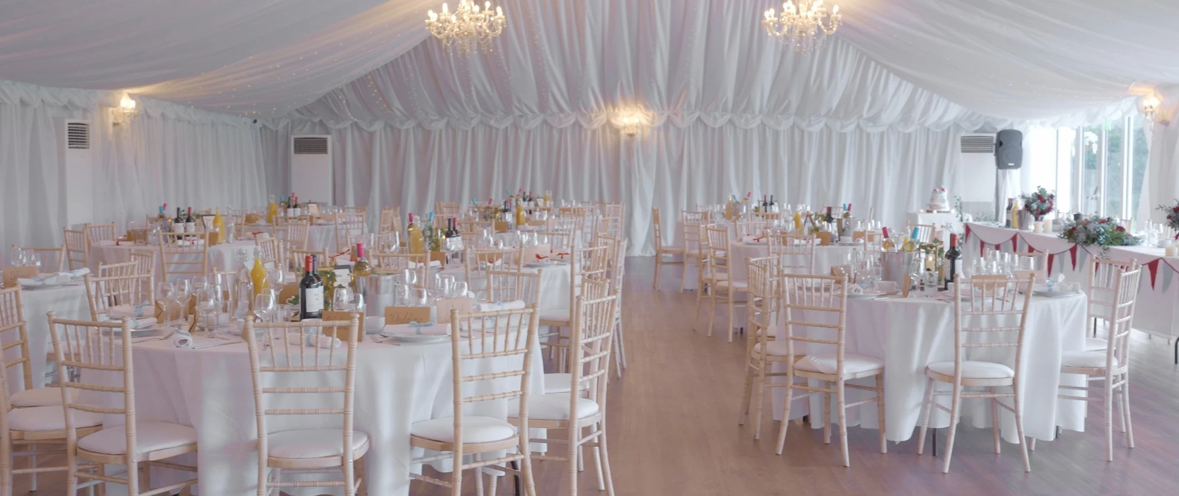 Elegant event space with round tables draped in white tablecloths, set with silverware, glasses, and wine bottles. Pale wood chairs surround the tables. The ceiling is decorated with white drapes and chandeliers, and the walls are covered with white 