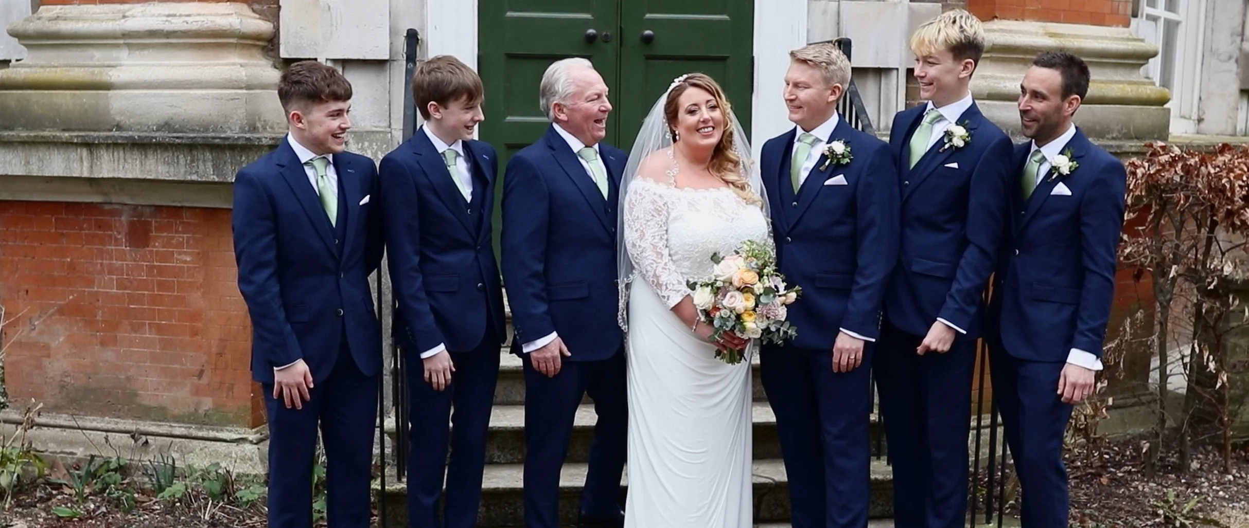 Group of wedding party members, including the bride in a white dress holding a bouquet, and six men in matching blue suits with light green ties, standing outside in front of a brick building with green doors, smiling and laughing together.