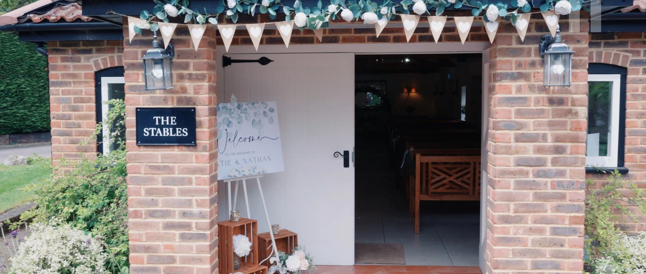 Brick building entrance decorated with white bunting, flowers, and lanterns for a wedding celebration, with a sign welcoming guests to the wedding of Katie and Nathan.