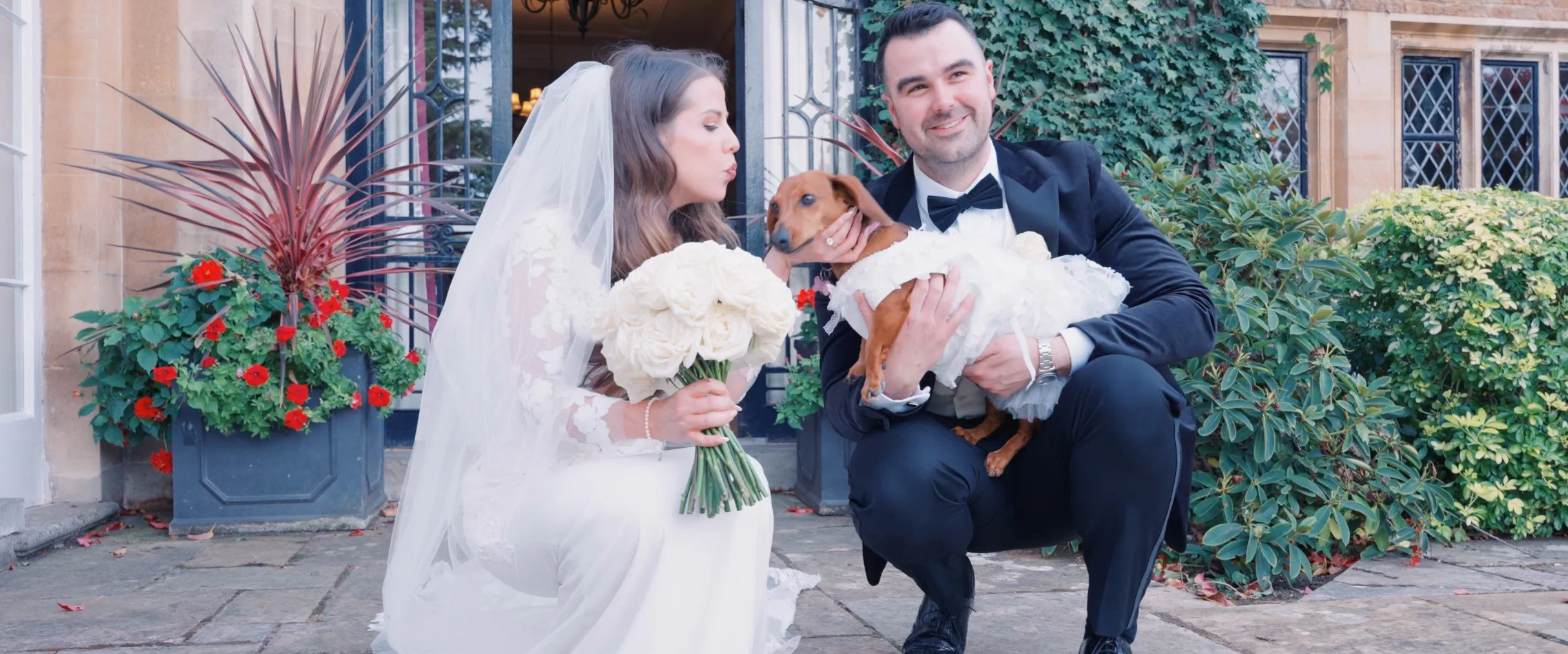 A couple in wedding attire, woman in a white wedding dress and veil, man in a tuxedo, posing with their dog dressed in a white dress at the entrance of a building with greenery and red flowers around.