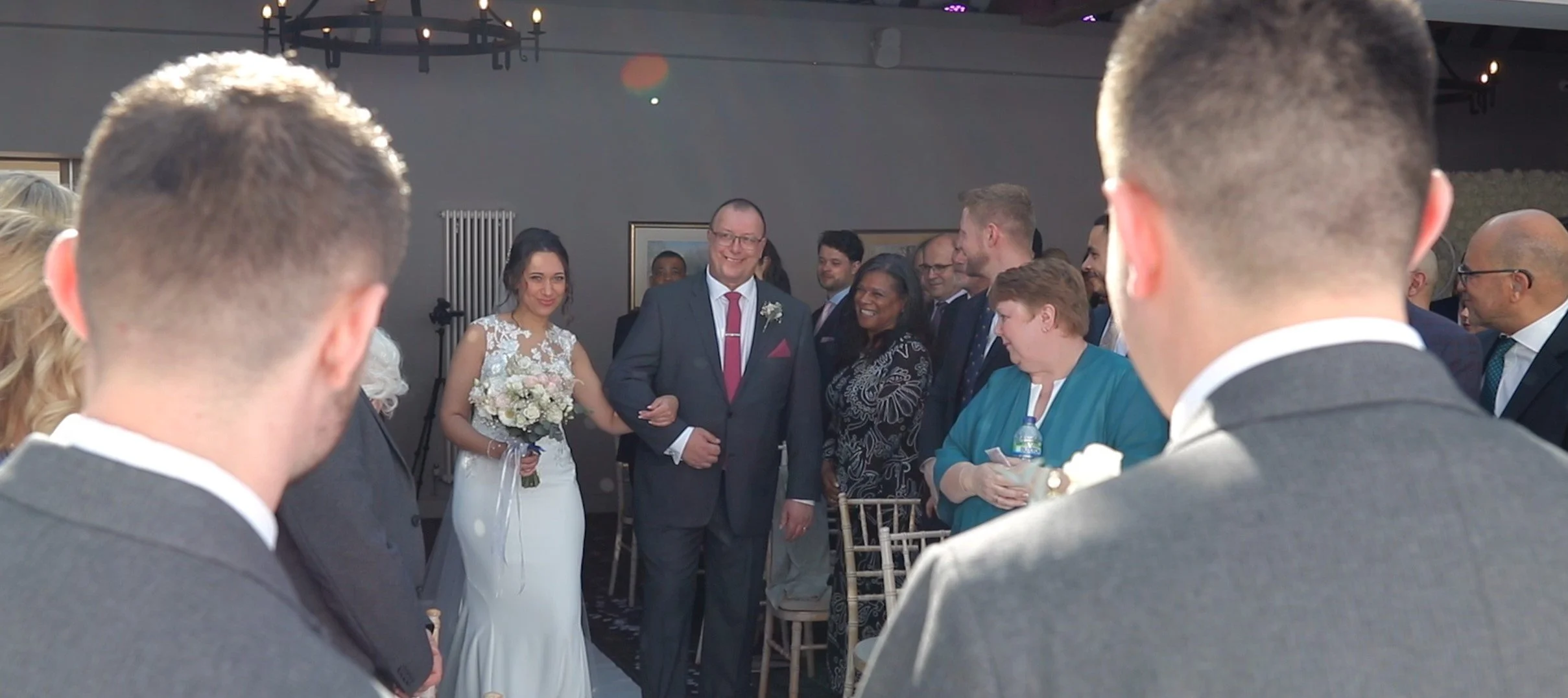 A wedding ceremony with the bride walking down the aisle, accompanied by a man in a suit, surrounded by guests who are smiling and seated, indoors with dim lighting.