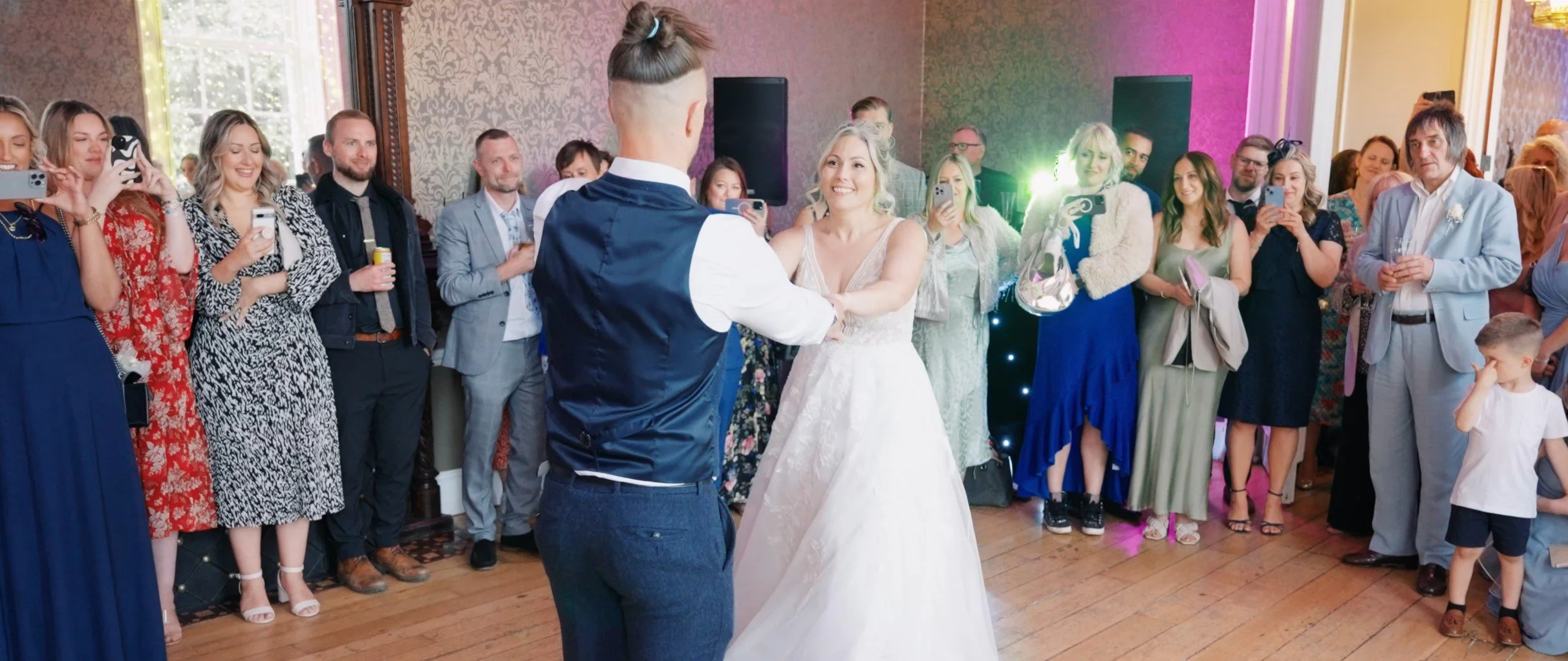 A bride and groom holding hands and looking at each other during their wedding dance, surrounded by guests taking photos and watching in a decorated indoor venue.