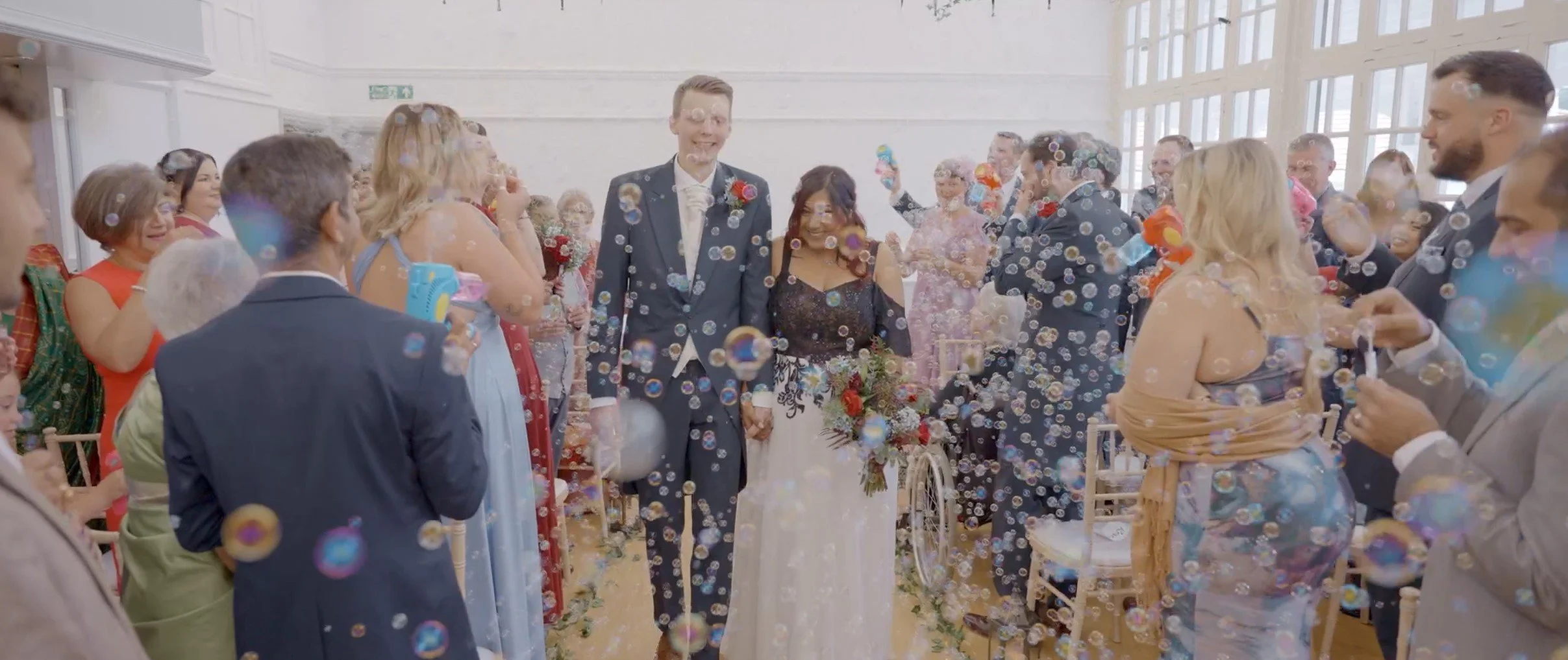 A joyful wedding celebration with a bride and groom walking hand in hand through a shower of bubbles, surrounded by friends and family in a bright indoor venue.