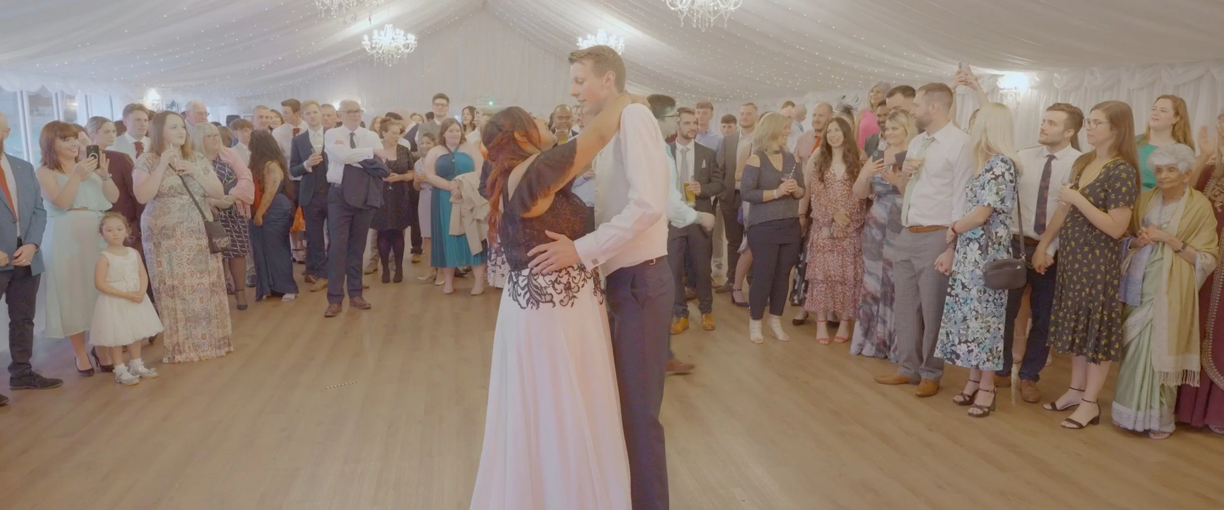 A couple is dancing in the center of a reception hall, surrounded by a crowd of guests watching and taking photos at a wedding celebration.