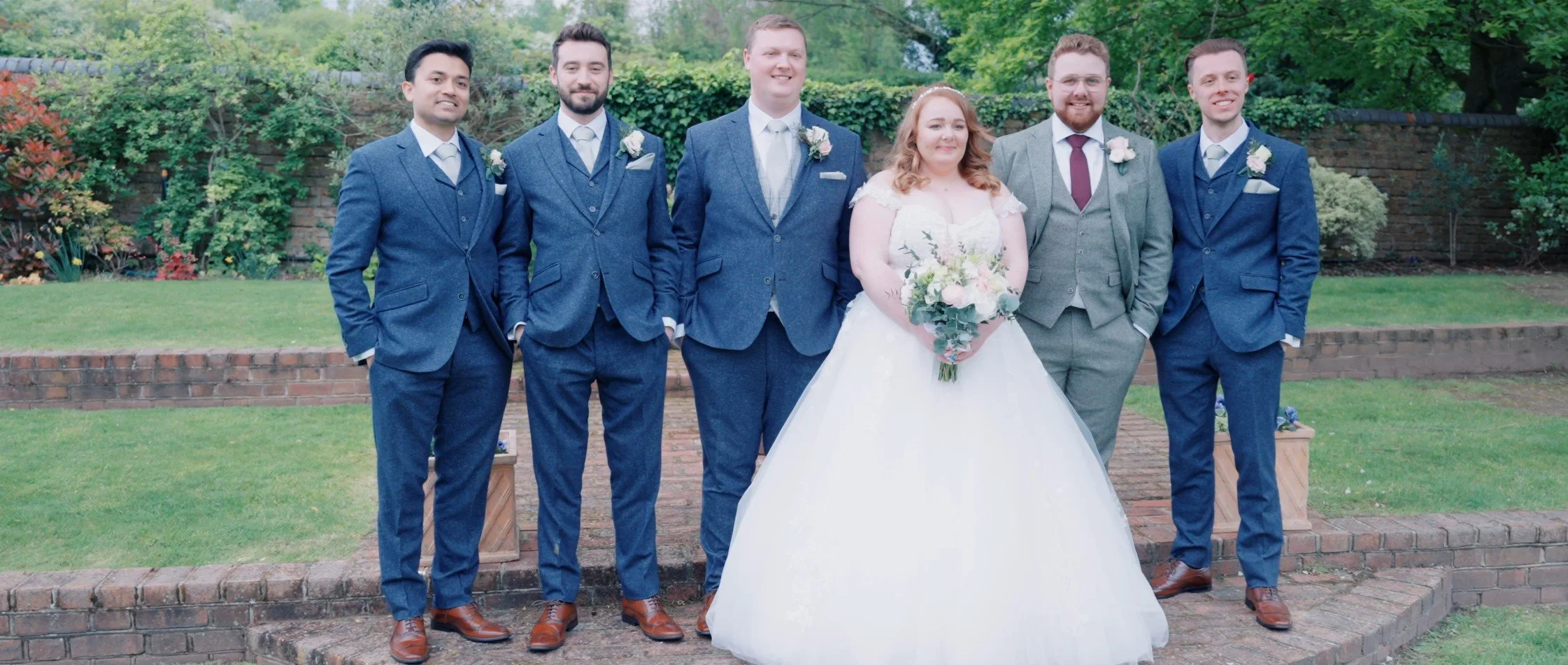 Group of six people, including a bride in a white wedding dress holding a bouquet, and five men in blue and grey suits, standing outdoors on a brick walkway with greenery and trees in the background.