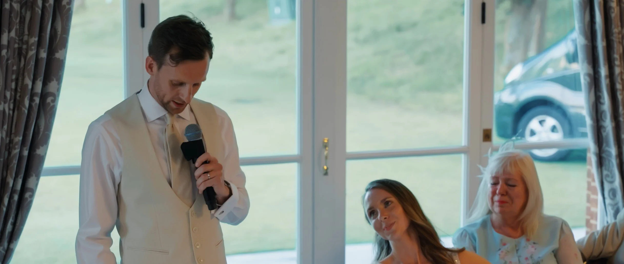 Man in white vest and shirt holding a microphone, wedding reception scene with two women sitting, one smiling and the other with eyes closed, near a window with curtains, outside view of grass and a car.