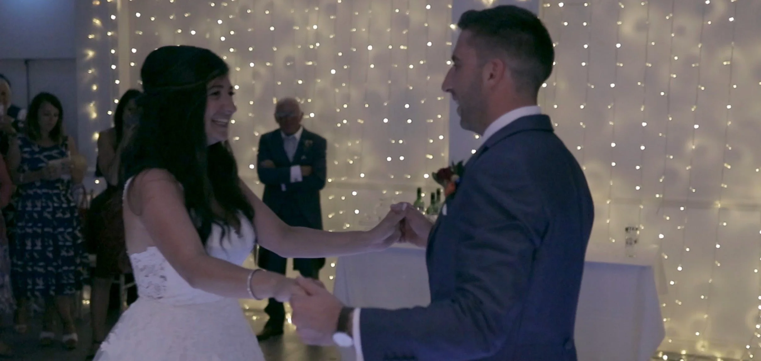 A bride and groom dancing at their wedding reception, holding hands and smiling, with guests and string lights in the background.
