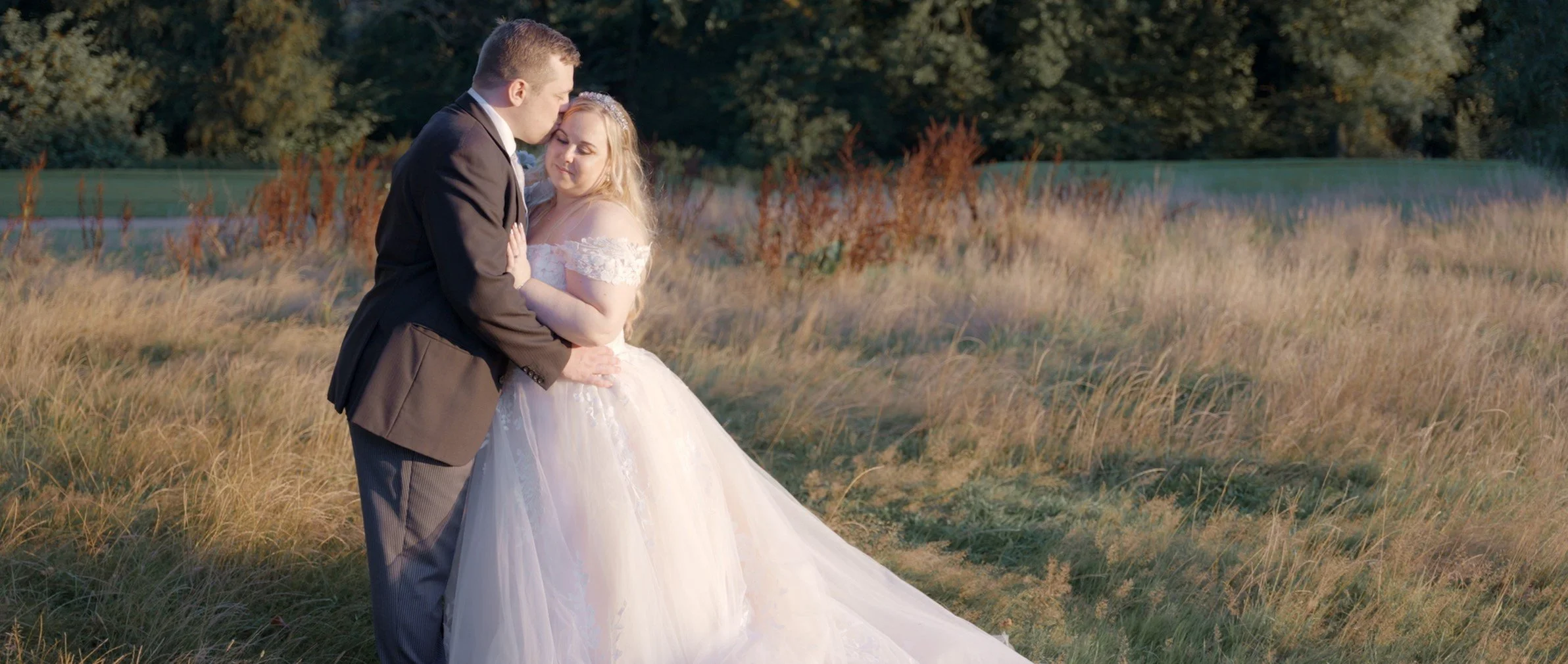 A bride and groom sharing an intimate moment in a grassy outdoor field during sunset, with the groom leaning in for a kiss and the bride smiling softly.