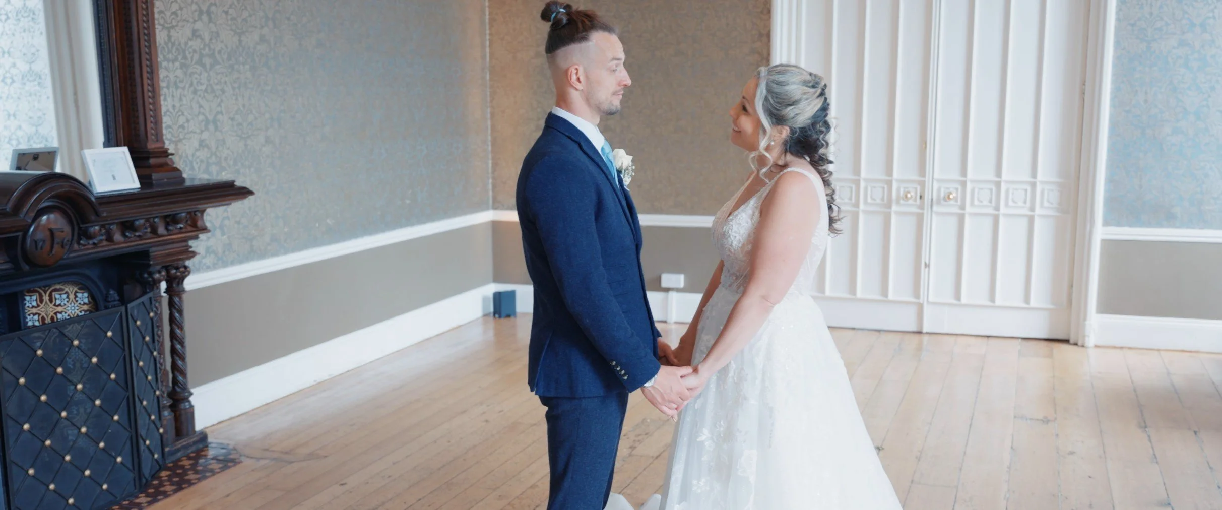 A bride and groom holding hands during their wedding, standing face to face indoors in a room with wooden floors and ornate decor.
