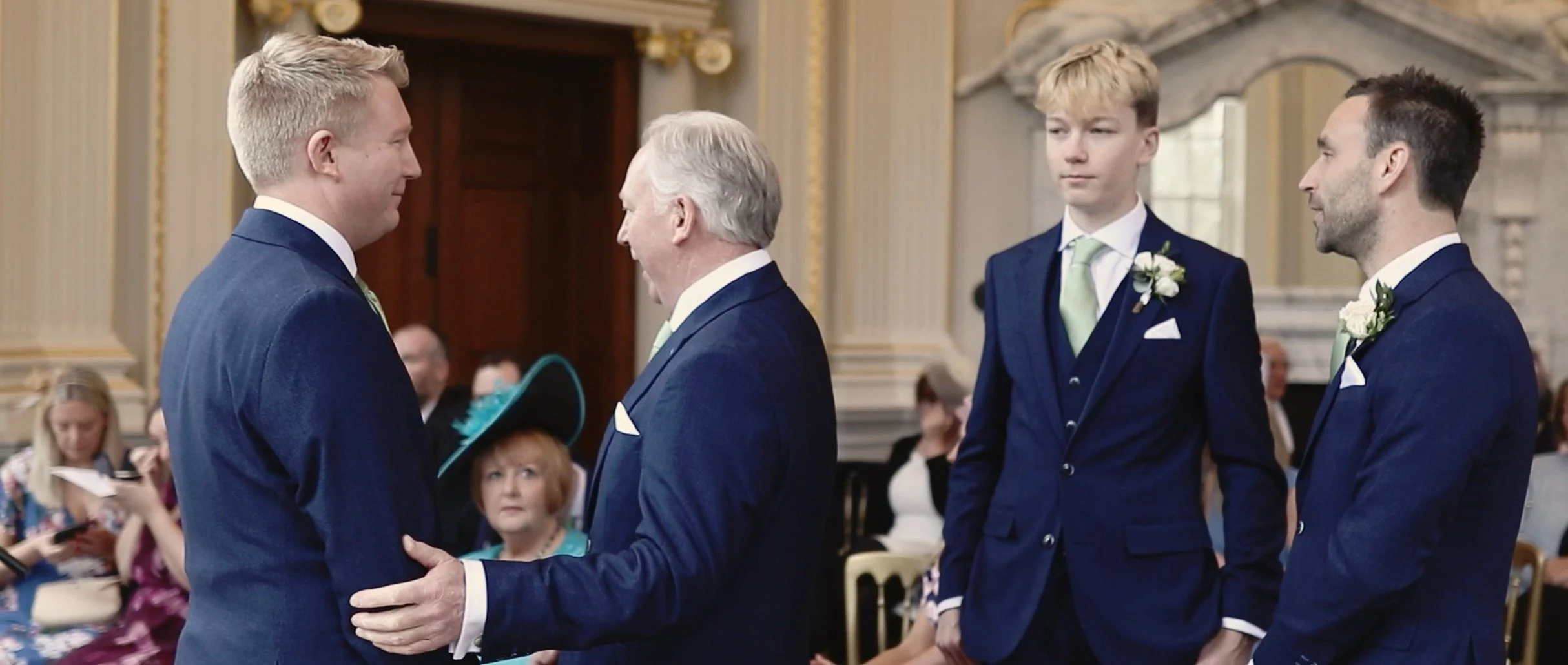 A wedding ceremony with four men dressed in blue suits, exchanging vows in an ornate room with gold accents. Two women are seated in the background, one using a phone and the other reading, while a red-haired woman wearing a teal hat observes.