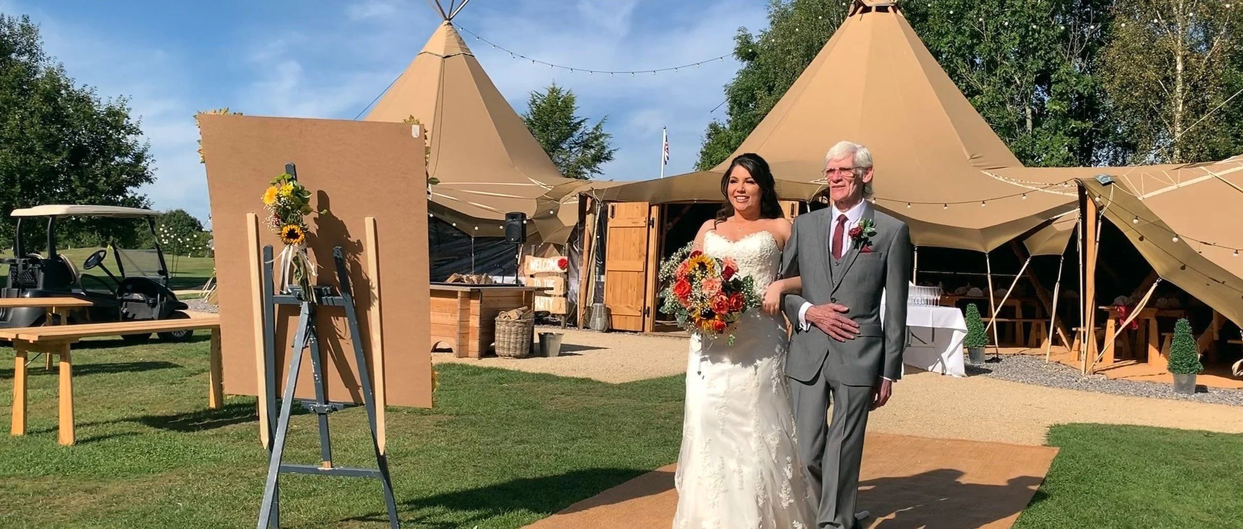 A bride dressed in a white wedding gown holding a colorful bouquet walking arm-in-arm with an elderly man in a gray suit at an outdoor wedding venue with beige tents, trees, and grass on a sunny day.