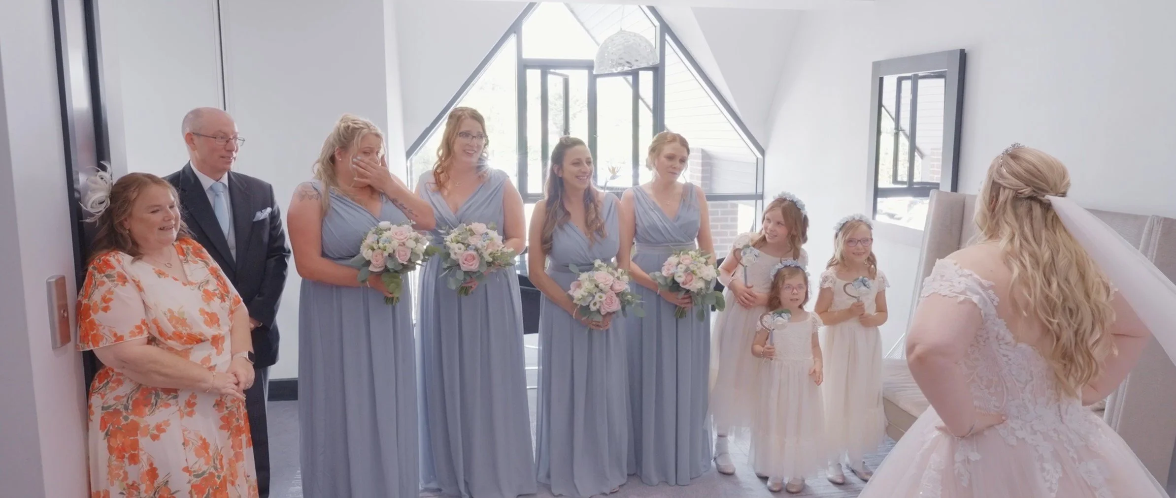 Bride with long blonde hair in a wedding dress stands with her back to the camera, facing a group of bridesmaids, flower girls, and family members in a bright, modern room with large windows.