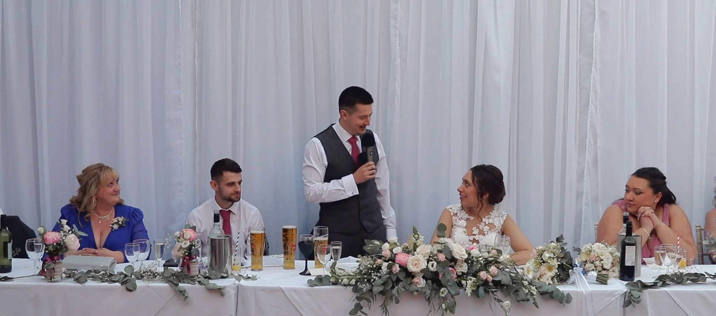 A wedding reception scene with five people sitting at a decorated head table. The man in the center is giving a speech, holding a microphone, and smiling at the woman in a white dress. The women and men are dressed formally, with the woman on the far