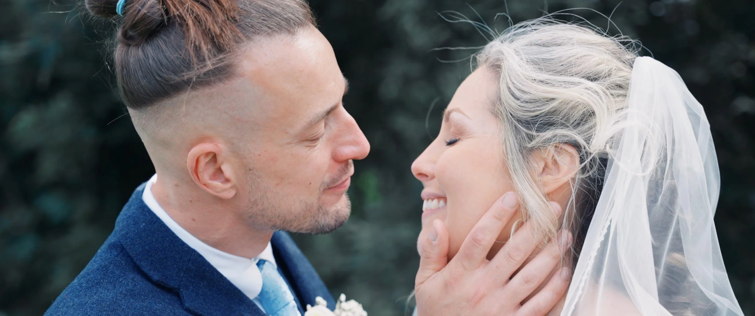 A bride and groom on their wedding day, close together with eyes closed, smiling, outdoors.