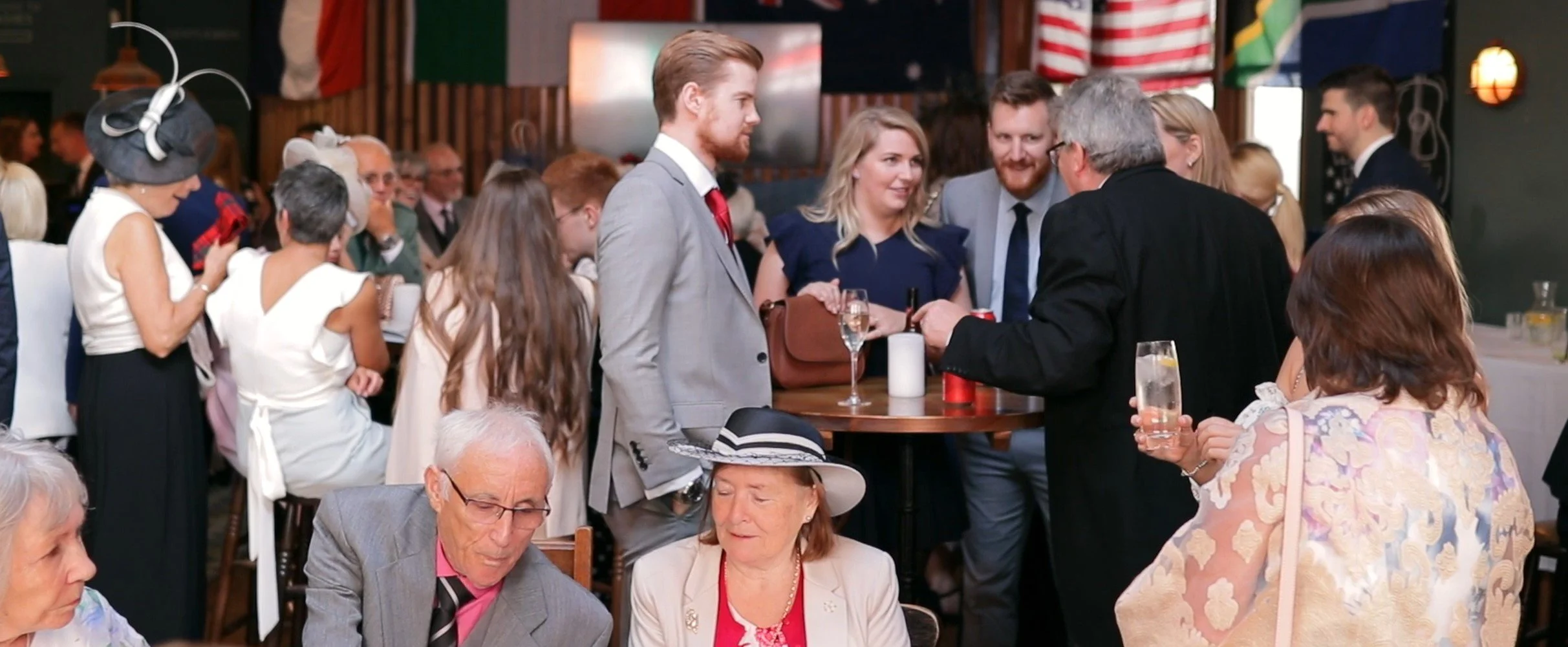Group of people at a social gathering, some dressed in formal attire, in a decorated indoor setting with flags and string lights.