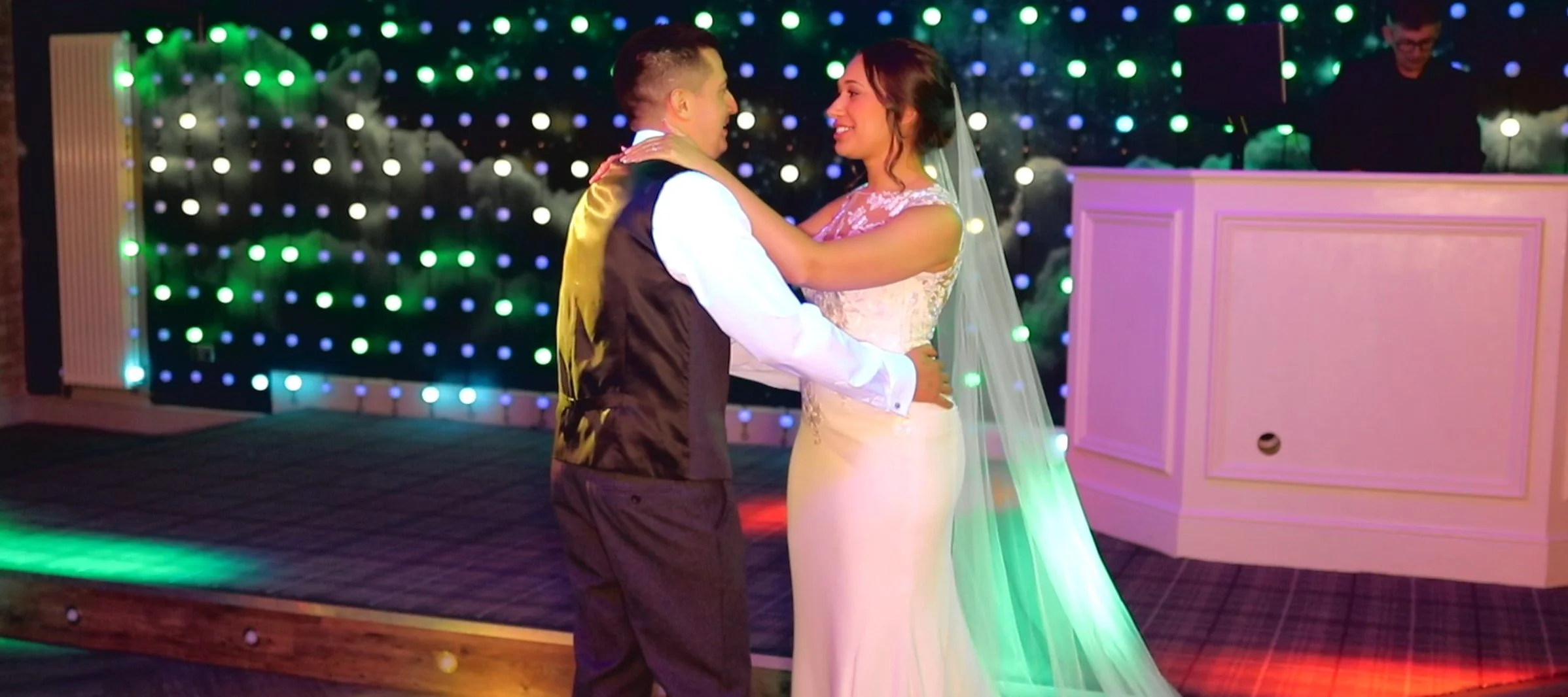 A bride and groom share a dance at their wedding reception with colorful lights in the background.