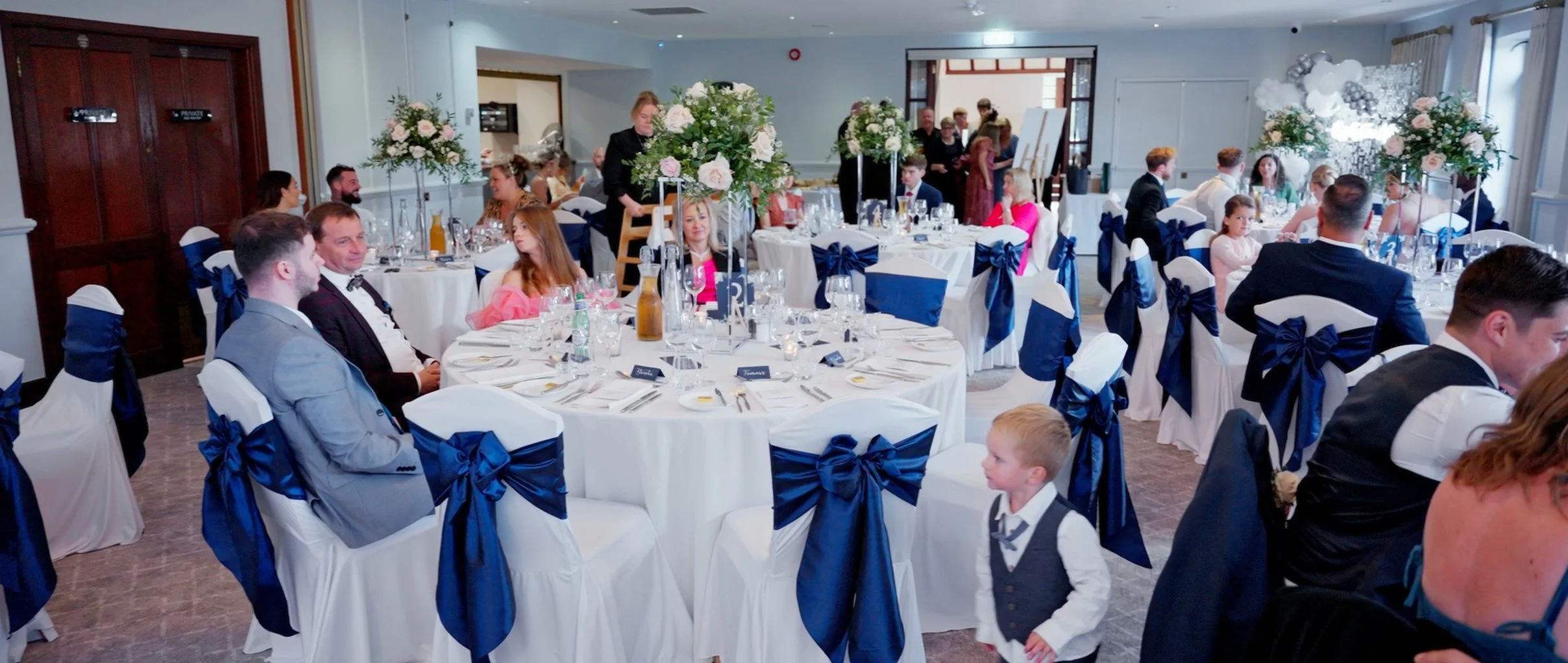 Guests seated at decorated tables with white tablecloths, navy blue sashes, and floral centerpieces in a banquet hall during a celebration or wedding reception.