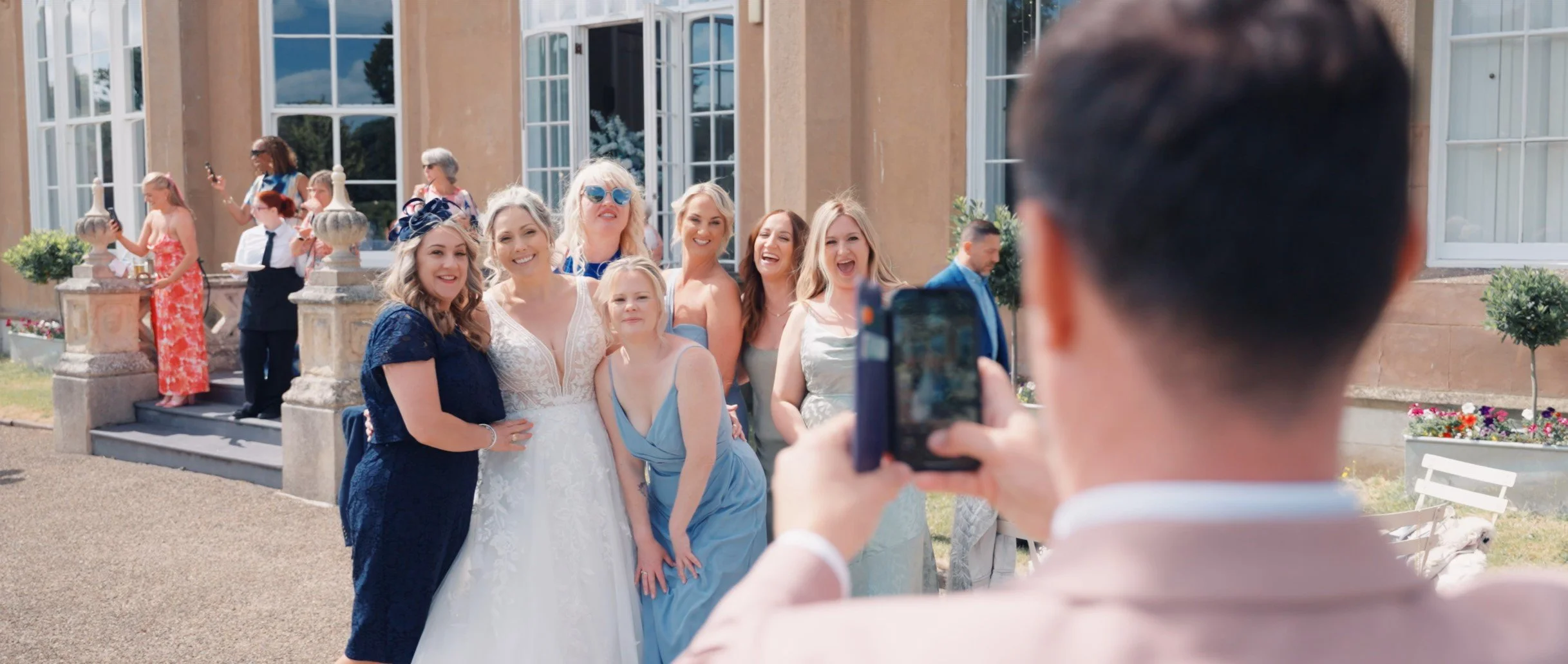 People taking a group photo outside a building, with a woman in a white wedding dress and others in colorful dresses, and a man in a suit in the foreground holding a phone.