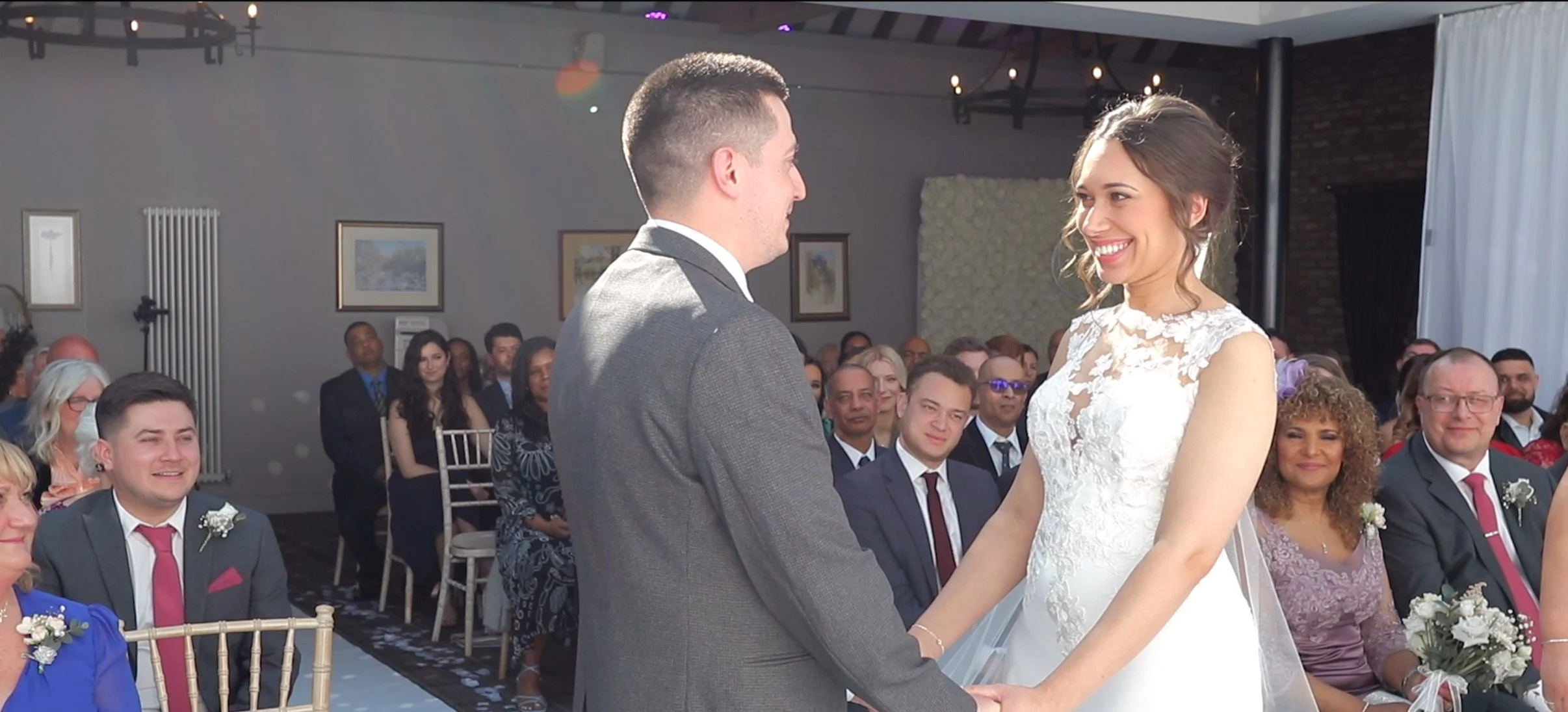 A bride and groom exchange vows at their wedding ceremony in a decorated indoor venue with seated guests watching.