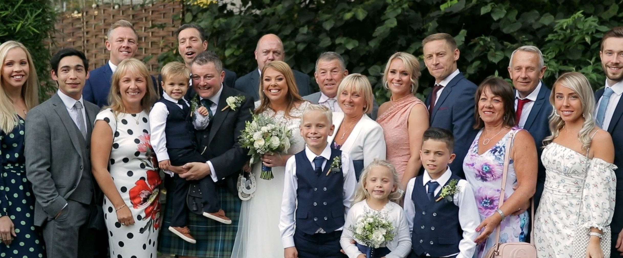 Group of people dressed in formal attire at a wedding. The bride is holding a bouquet, and the groom is in a tuxedo. Children and adults are smiling for the photo outdoors, with greenery in the background.