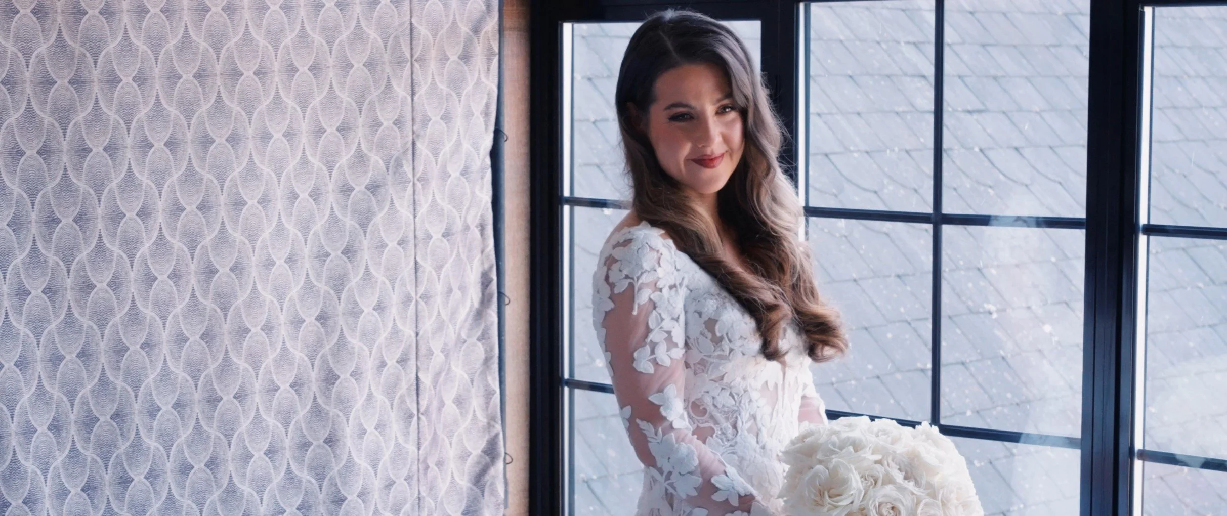 A woman in a white dress with floral patterns stands by a window holding a bouquet of white roses, smiling at the camera.