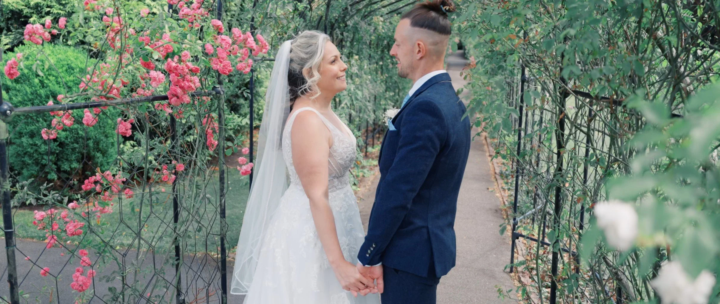A bride and groom holding hands and smiling at each other on a garden pathway surrounded by pink and white flowers and greenery.