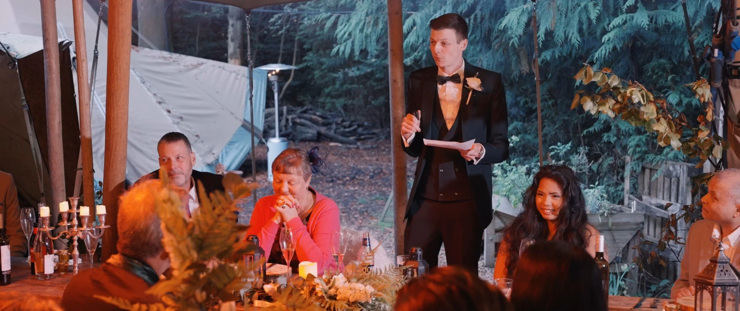 A man in a tuxedo giving a speech at a wedding reception seated at a decorated table, surrounded by guests, with outdoor tents and green trees in the background.
