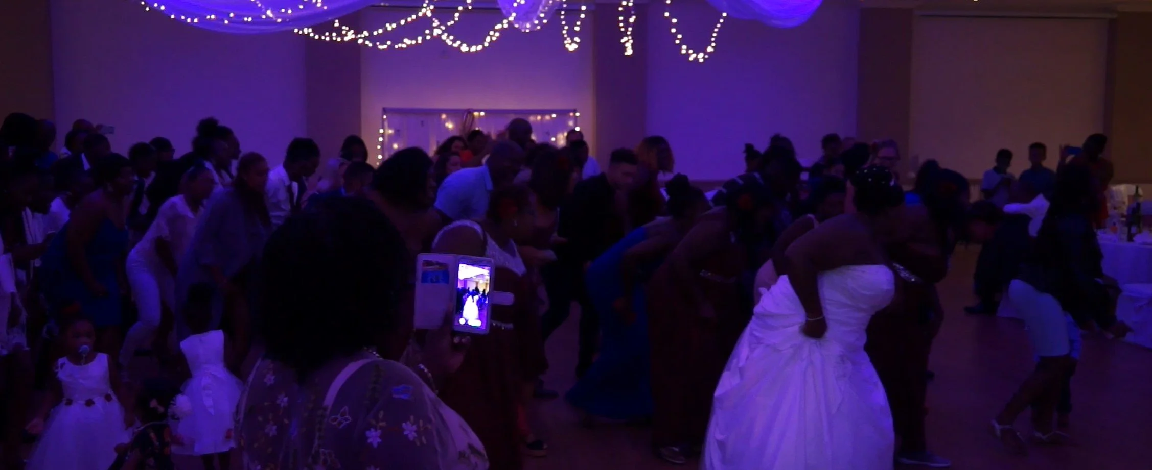 People dance at a party in a dimly lit room decorated with string lights. Many guests are dressed in formal attire, and some are capturing the moment with phones. A bride in a white gown is visible among the crowd.