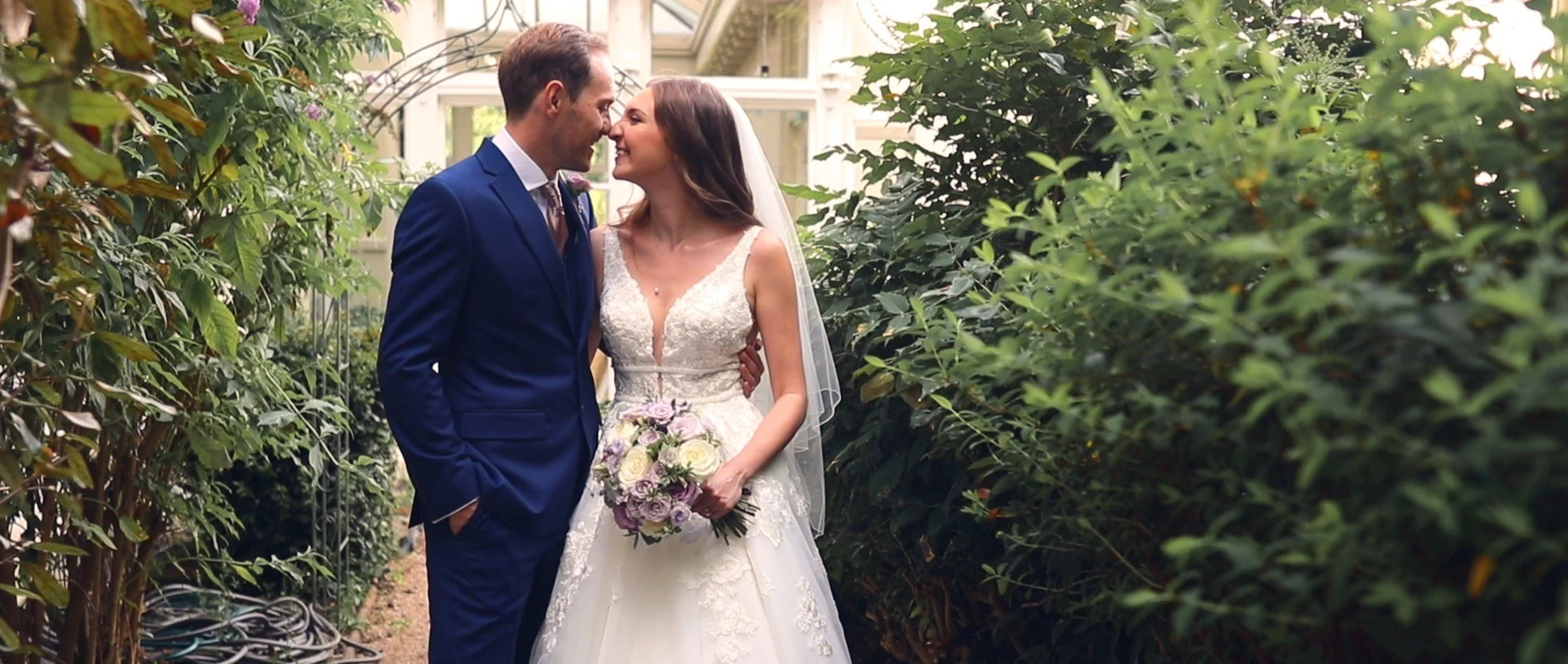 A bride and groom share a close moment on their wedding day, standing amidst lush greenery in a garden setting. The bride wears a lace wedding gown and holds a bouquet of white and light purple flowers. The groom is dressed in a navy blue suit.