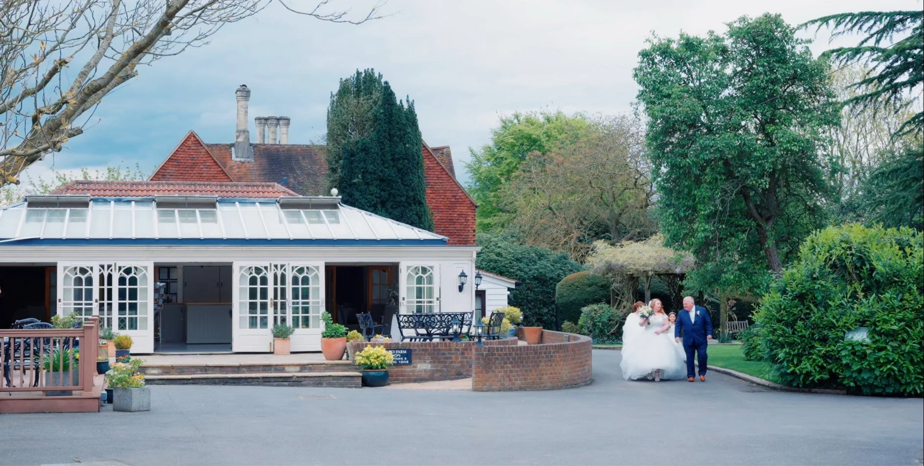 A bride and groom walking together outside a venue surrounded by trees and greenery, with the bride wearing a white wedding gown and holding a bouquet, and the groom in a dark suit.