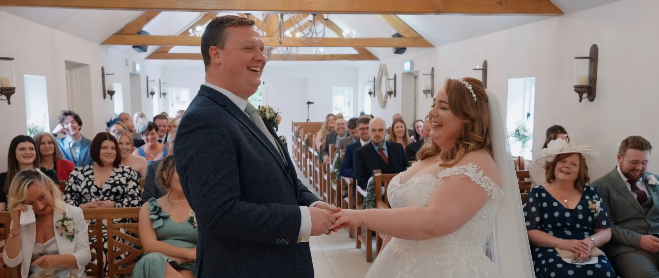 Bride and groom exchanging vows during wedding ceremony in a rustic chapel, guests seated in wooden pews watching and smiling.