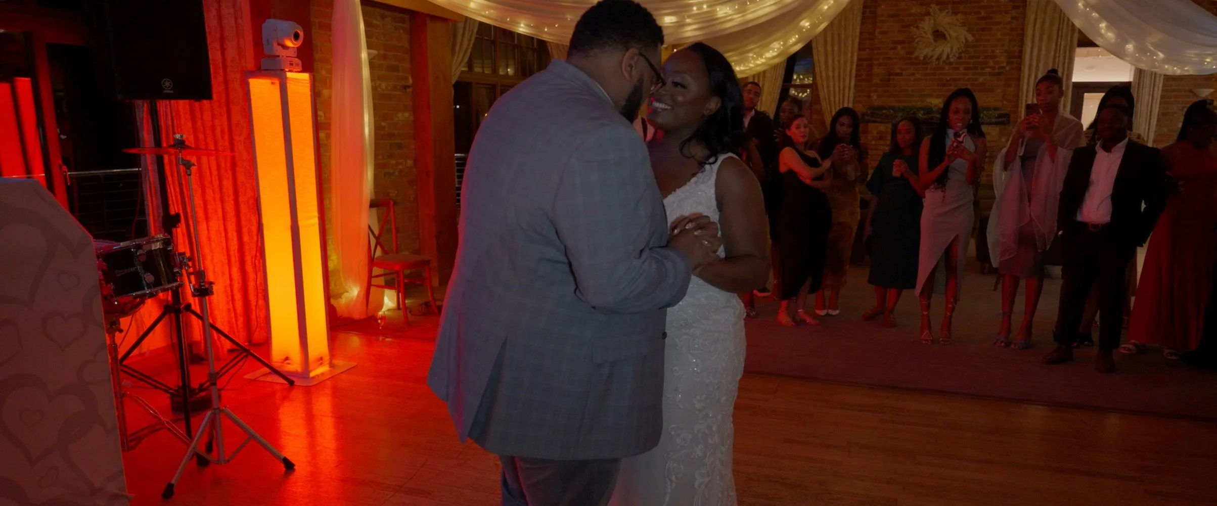A couple is dancing closely during a wedding reception, with guests watching and taking photos in the background, in a decorated indoor venue.