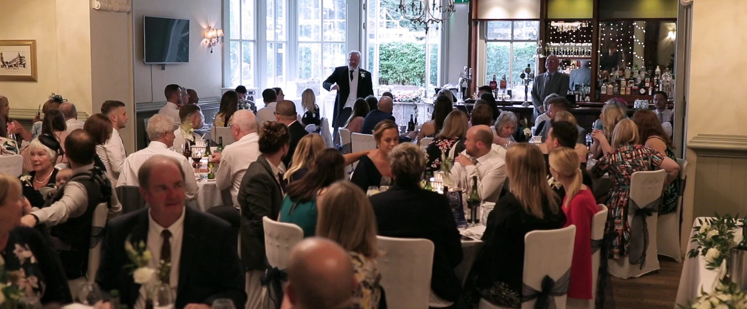 A wedding reception in a decorated indoor venue with guests seated at tables. A man in a tuxedo is speaking at a microphone near a large window. The room has soft lighting, floral arrangements, and a bar area in the background.