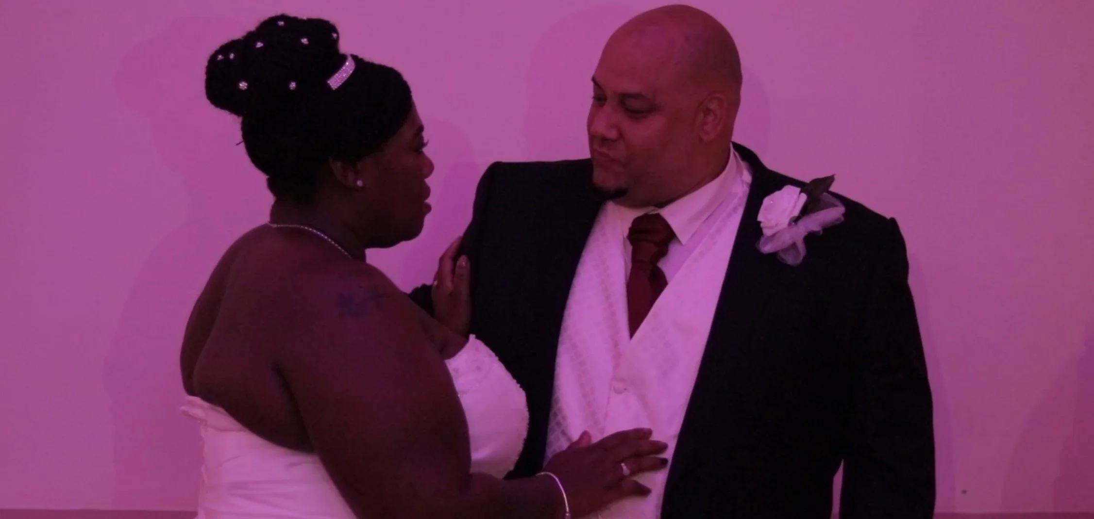 A couple dancing closely in formal attire, with the woman in a white dress and the man in a black suit with a boutonniere, sharing an intimate moment in a dimly lit room.
