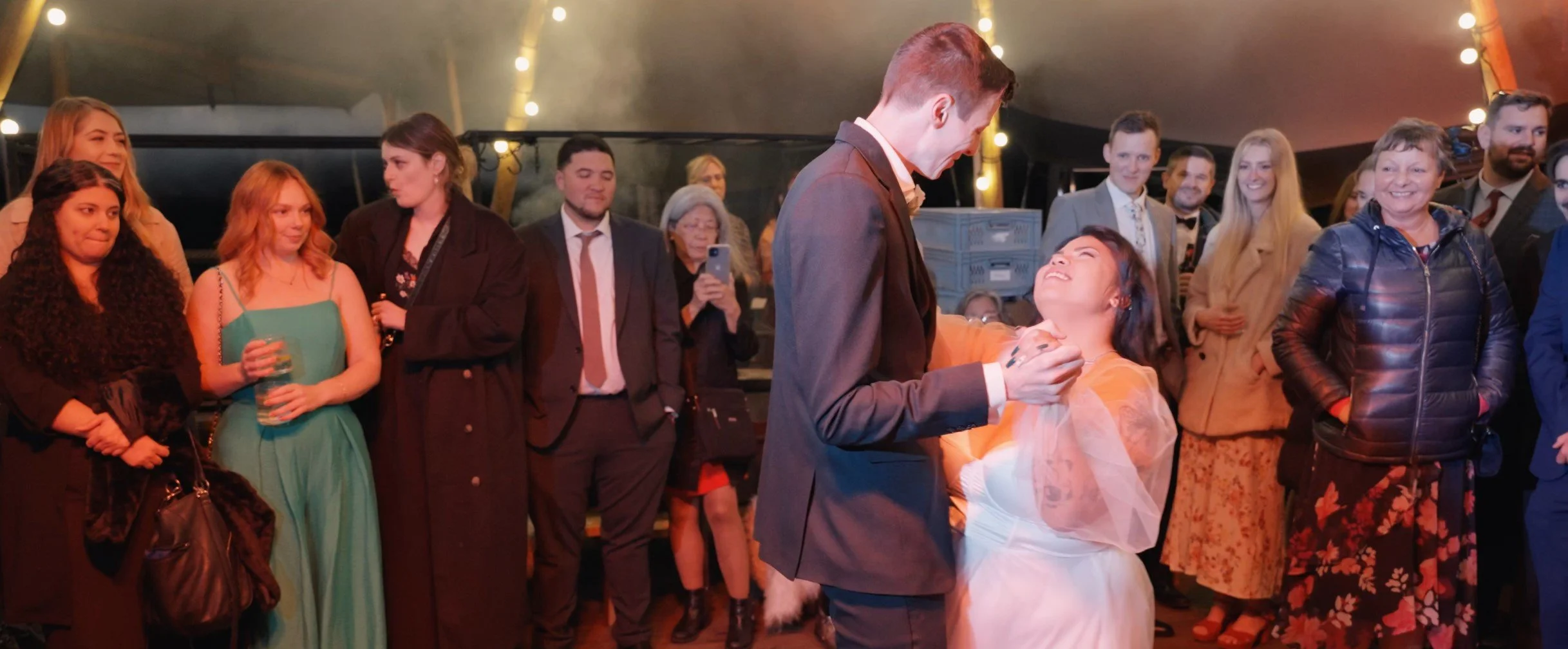 A wedding reception scene with a bride and groom dancing among guests. The groom is in a suit, and the bride is wearing a white dress with a veil. Guests are watching and smiling, some taking photos in a tent decorated with string lights.