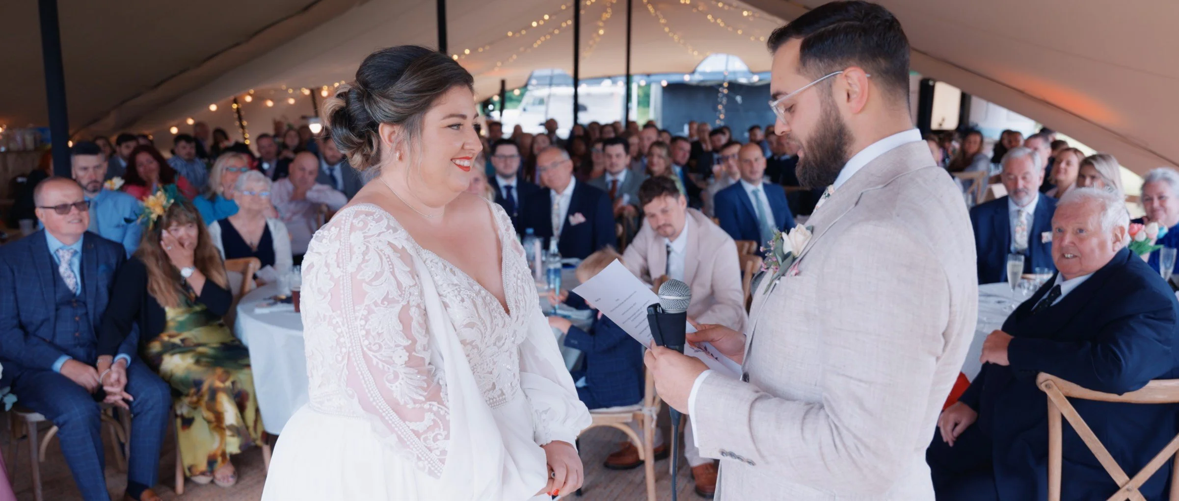 Bride and groom exchanging vows during a wedding ceremony under a decorated tent, with guests seated at tables watching.