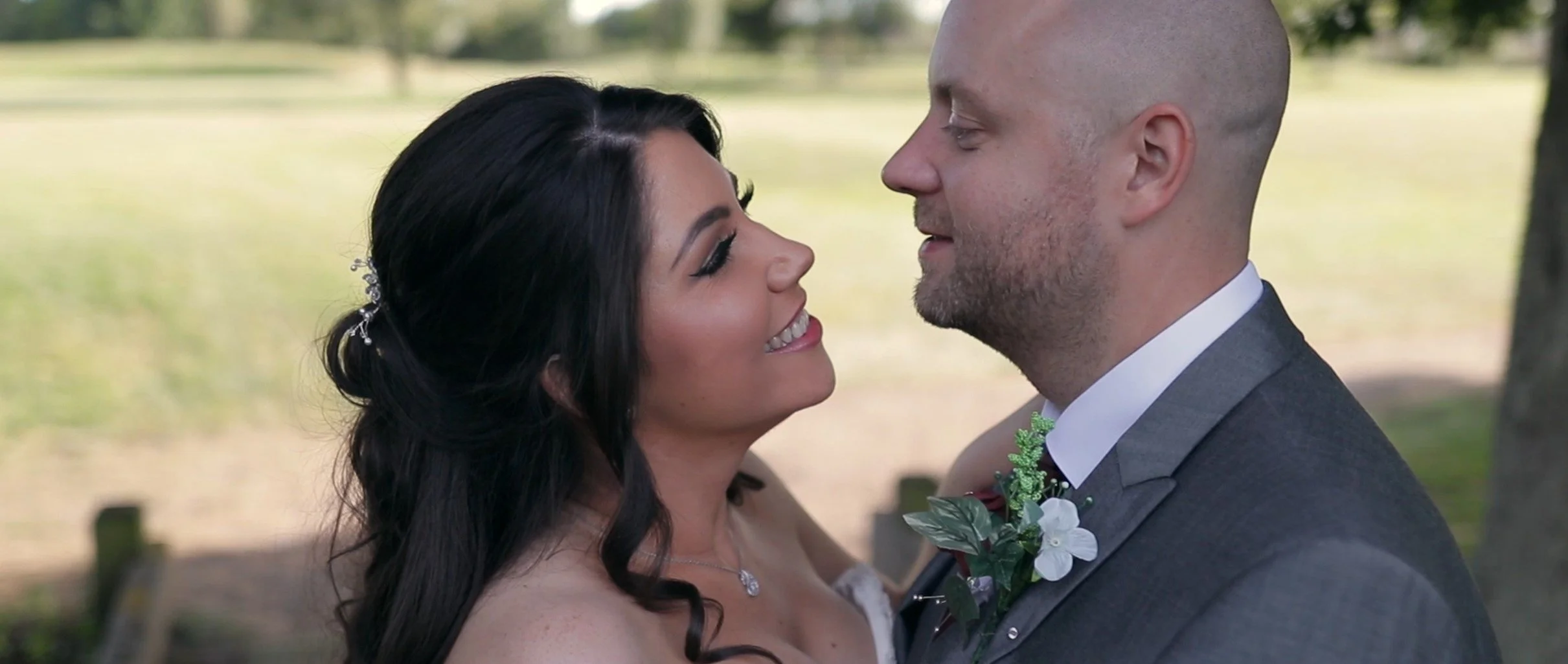 A bride and groom face each other outdoors during their wedding, with trees and a grassy area in the background, smiling and close.
