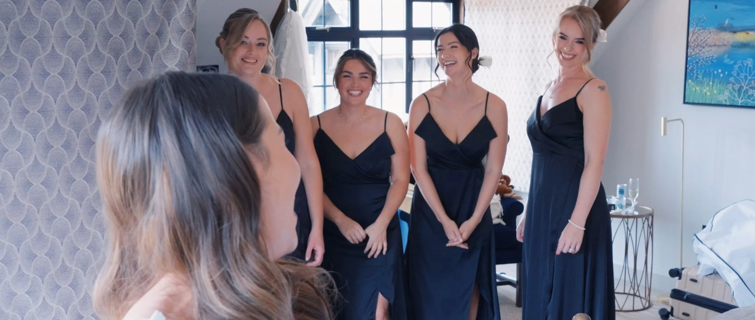 Group of five women in matching navy blue dresses, smiling and chatting in a bright room with large windows and modern decor.