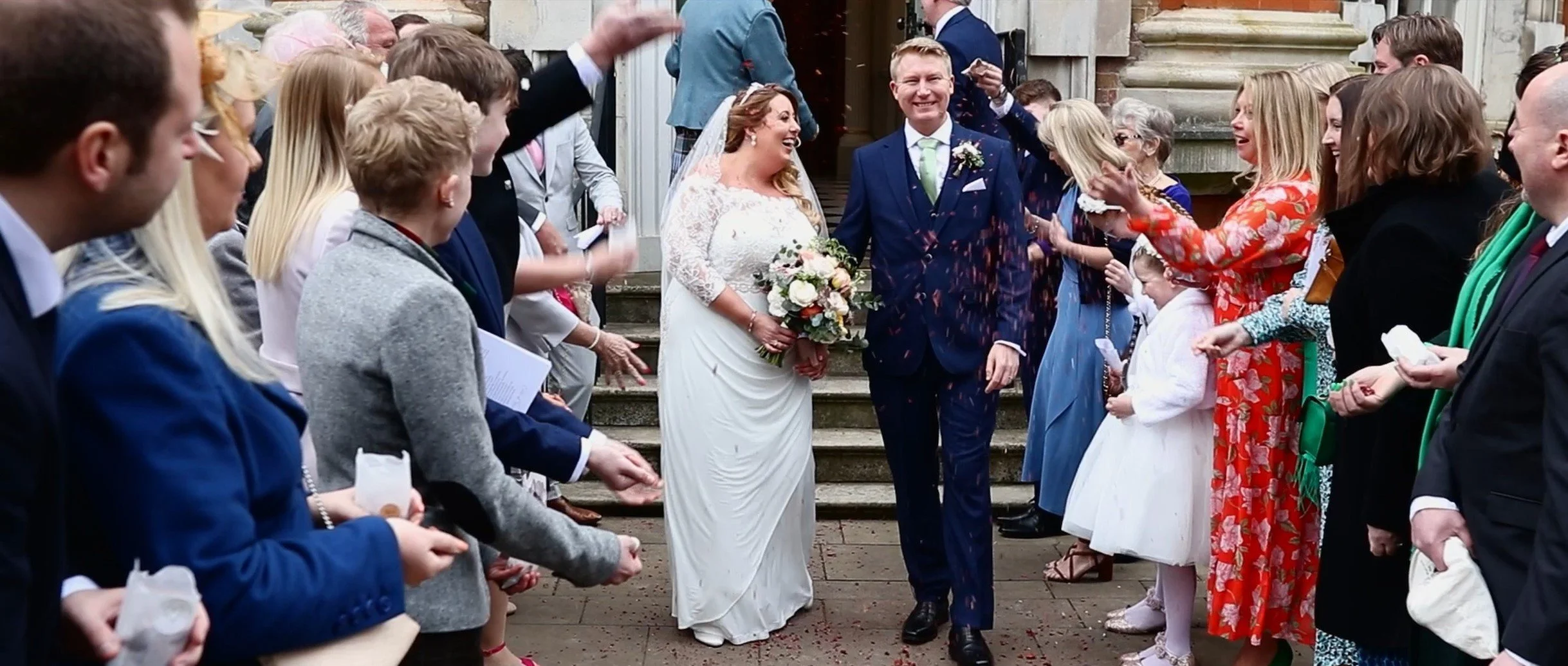 A wedding celebration with a bride in a white lace gown holding a bouquet, and a groom in a navy suit, walking down steps while guests throw confetti around them.